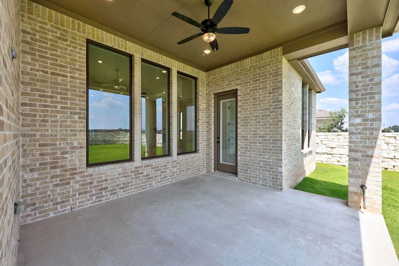 A covered patio with a concrete floor, brick walls, a ceiling fan, and a door leading inside, with a view of a grassy backyard and a stone fence.