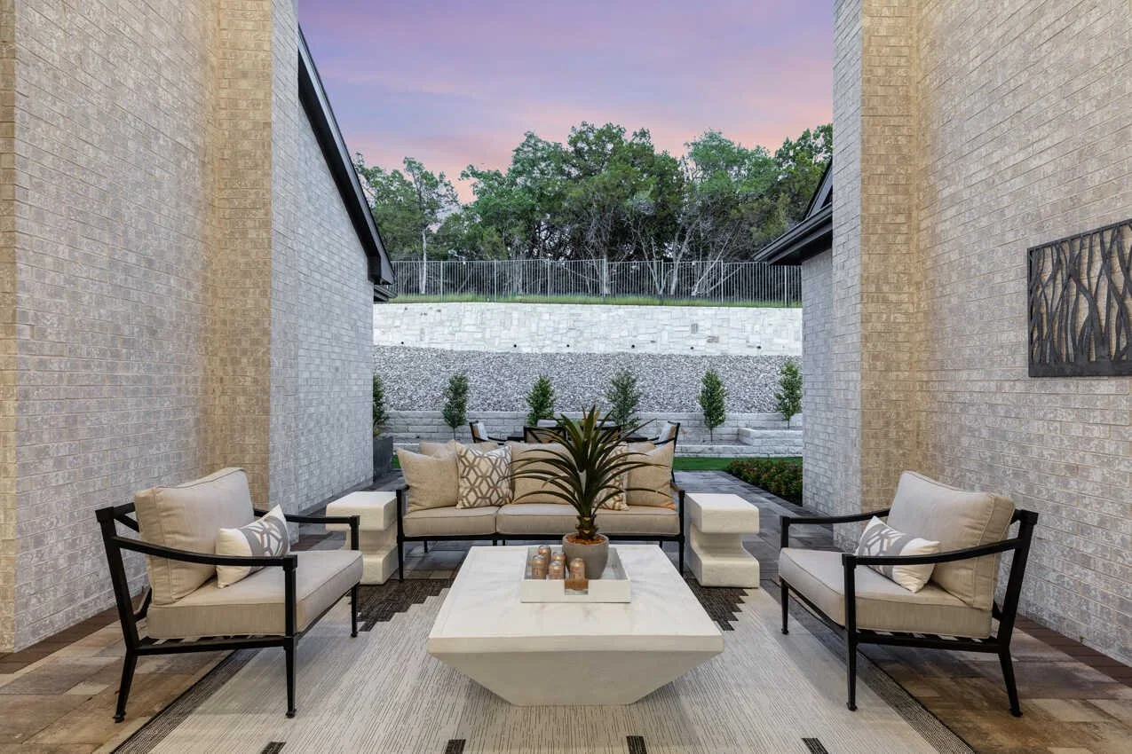 Modern outdoor patio with beige seating, black metal furniture, a central plant, and a sunset sky.