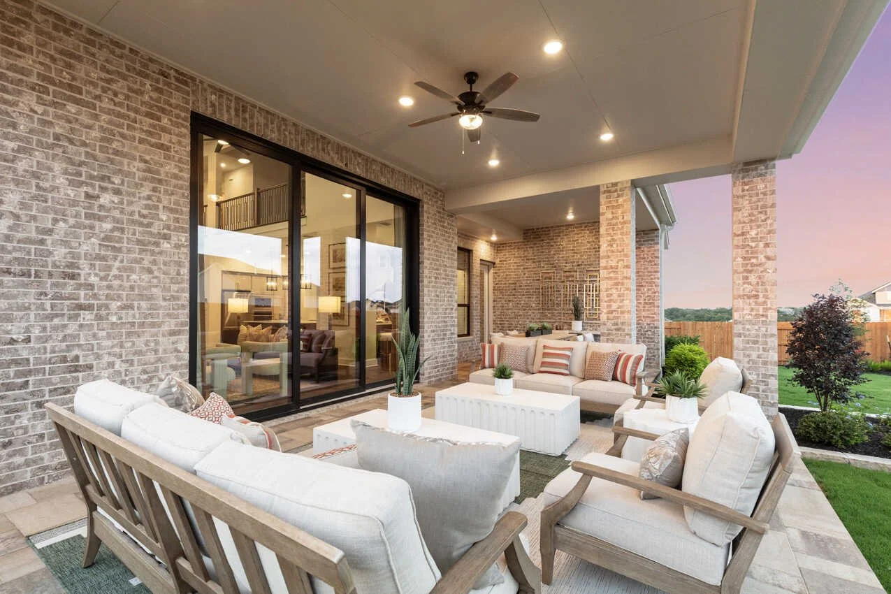 Covered outdoor patio with beige and white furniture, potted plants, a ceiling fan, brick walls, and sliding glass doors leading into a house at sunset.