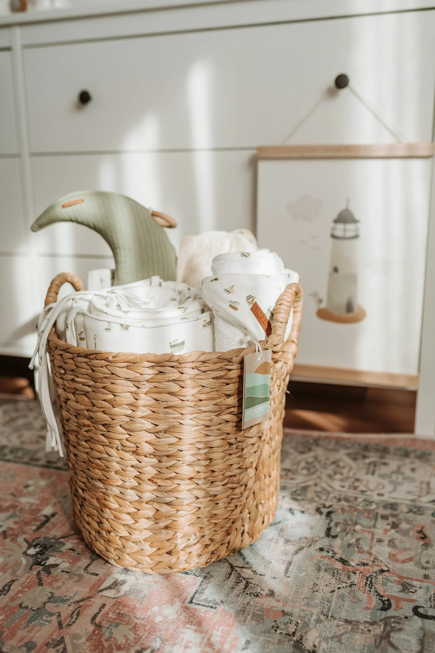 A woven basket containing white textiles and plush toys on a patterned rug in a room with a white dresser and a decorative lantern poster.