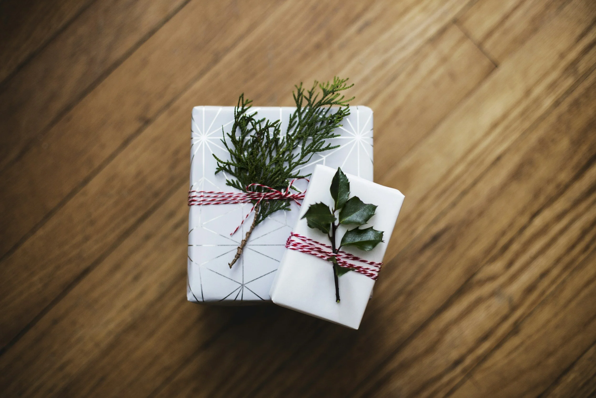 two boxes wrapped in white and silver wrapping paper, with red and white string and greenery, on top of a wooden table