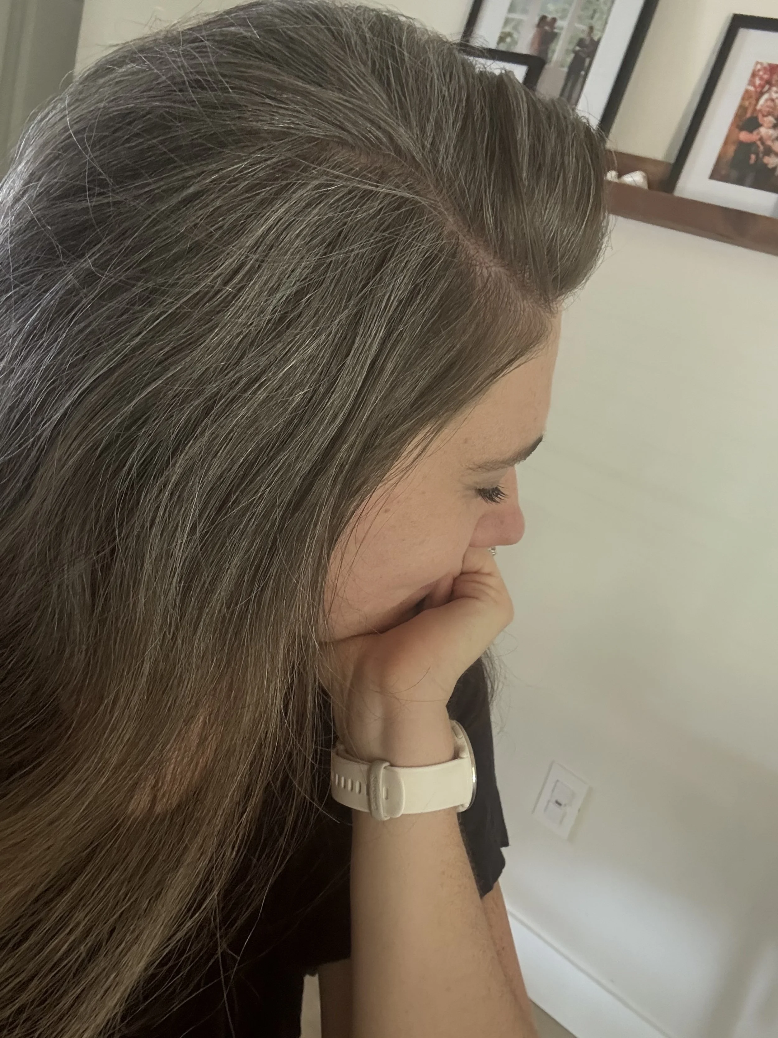 A woman with long, brown hair covering her face, resting her chin on her hand, wearing a white watch on her wrist, in a room with framed photos on the wall.