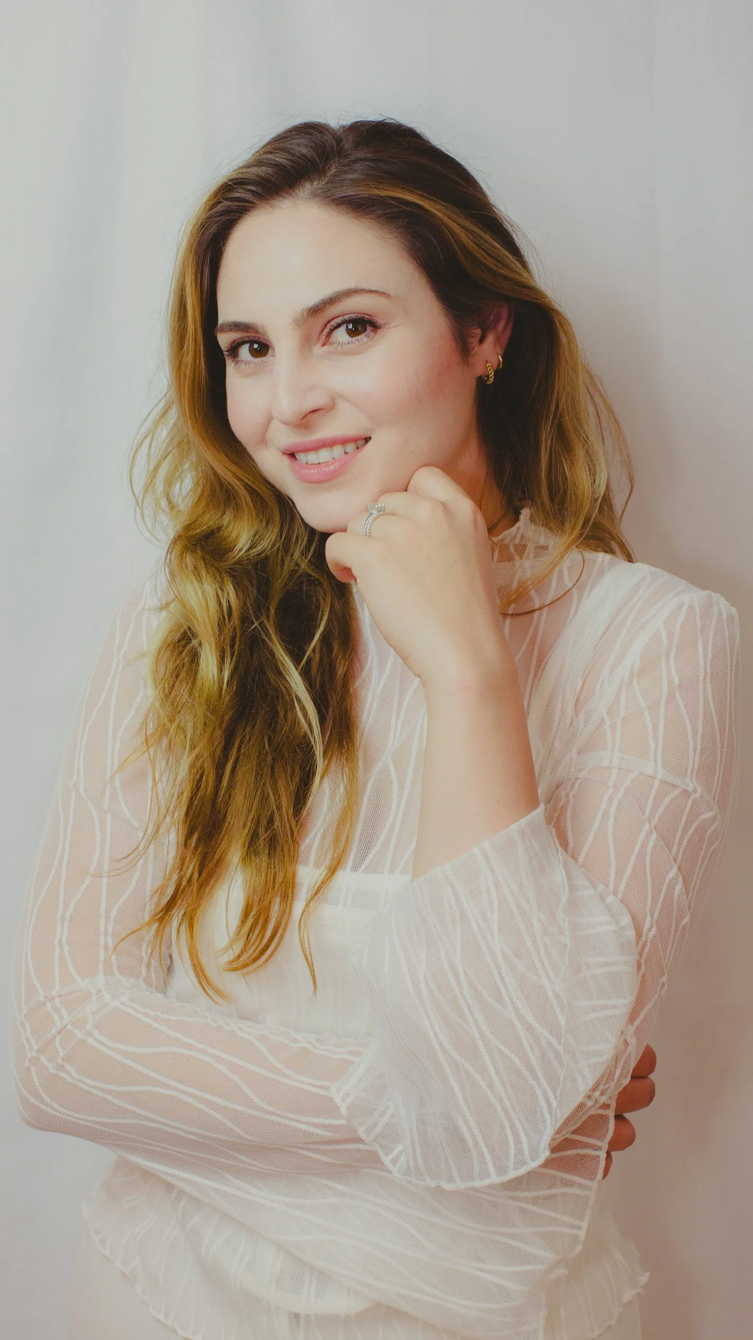 A young woman with wavy, shoulder-length hair, smiling and resting her chin on her hand, wearing a light-colored, patterned blouse, with earrings and a ring on her finger, standing against a plain wall.