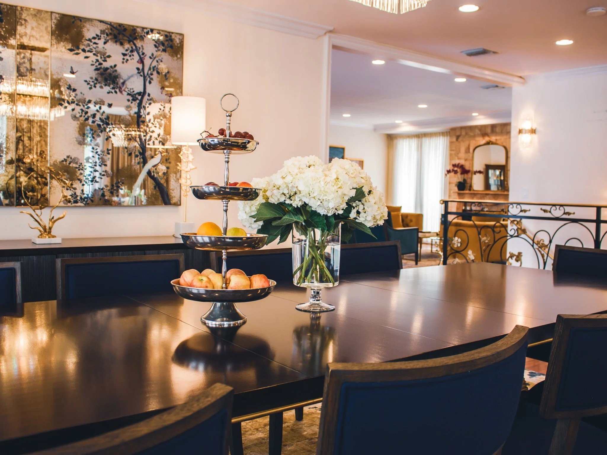 Elegant dining room with a large wooden table, flower centerpiece, and tiered fruit tray; modern decor, bright lighting, and elegant furniture.