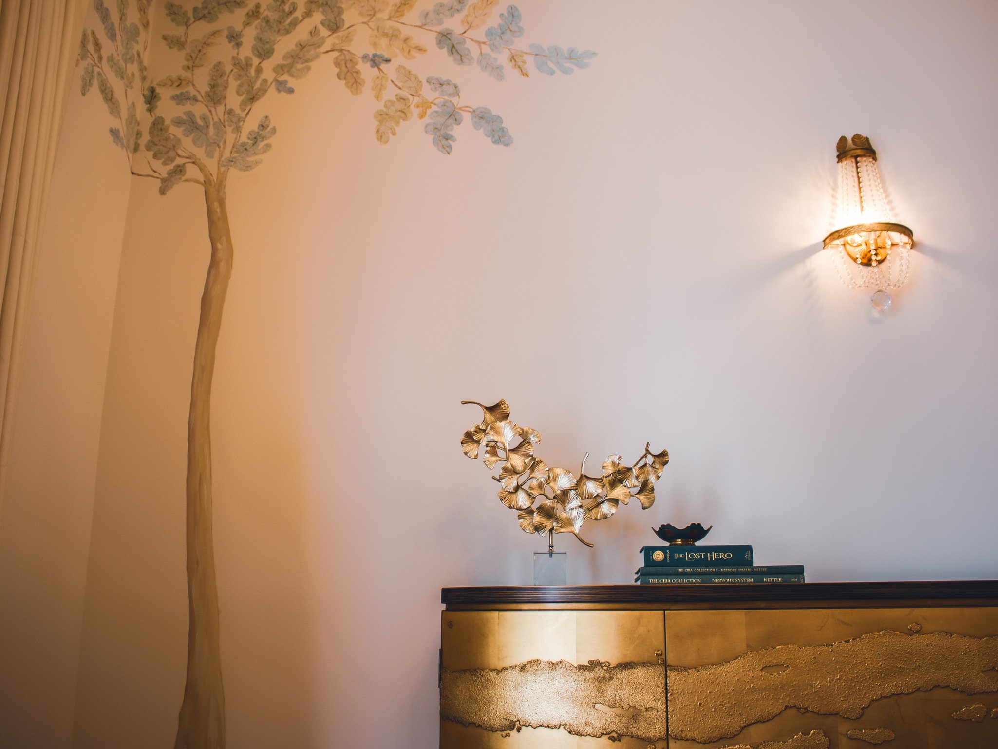 Interior wall decor featuring a wall-mounted sconce light, a tall painted tree on the wall, and a wooden cabinet with decorative objects and books on top.