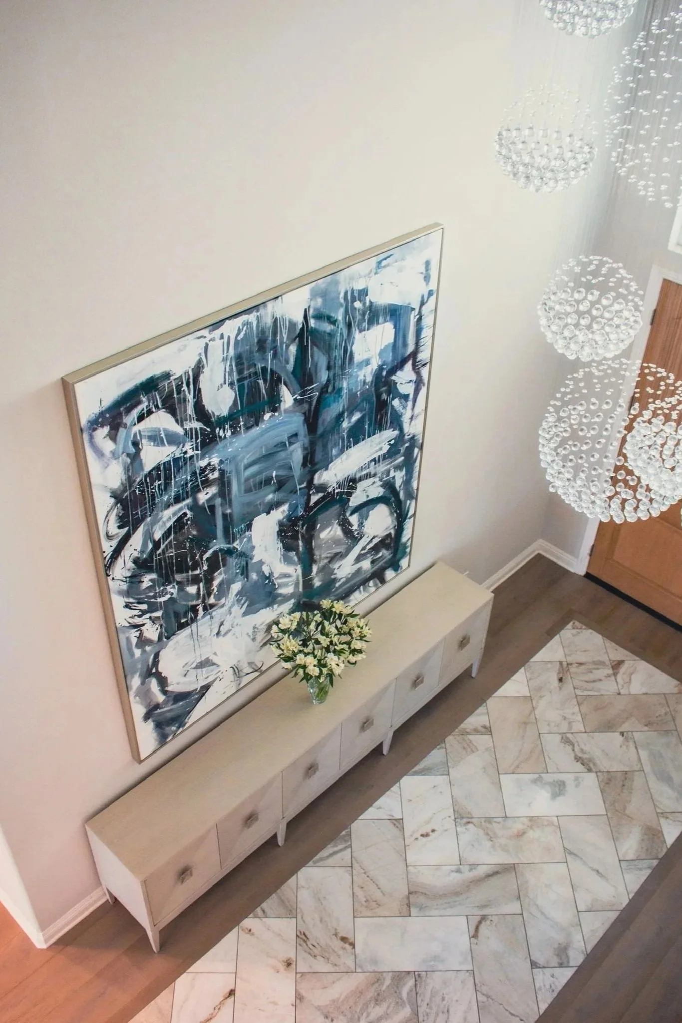 Overhead view of an entryway with a large abstract painting on a white wall, a white console with a vase of white flowers, a chandelier with spherical glass elements, and a tiled floor with a beige and white pattern.