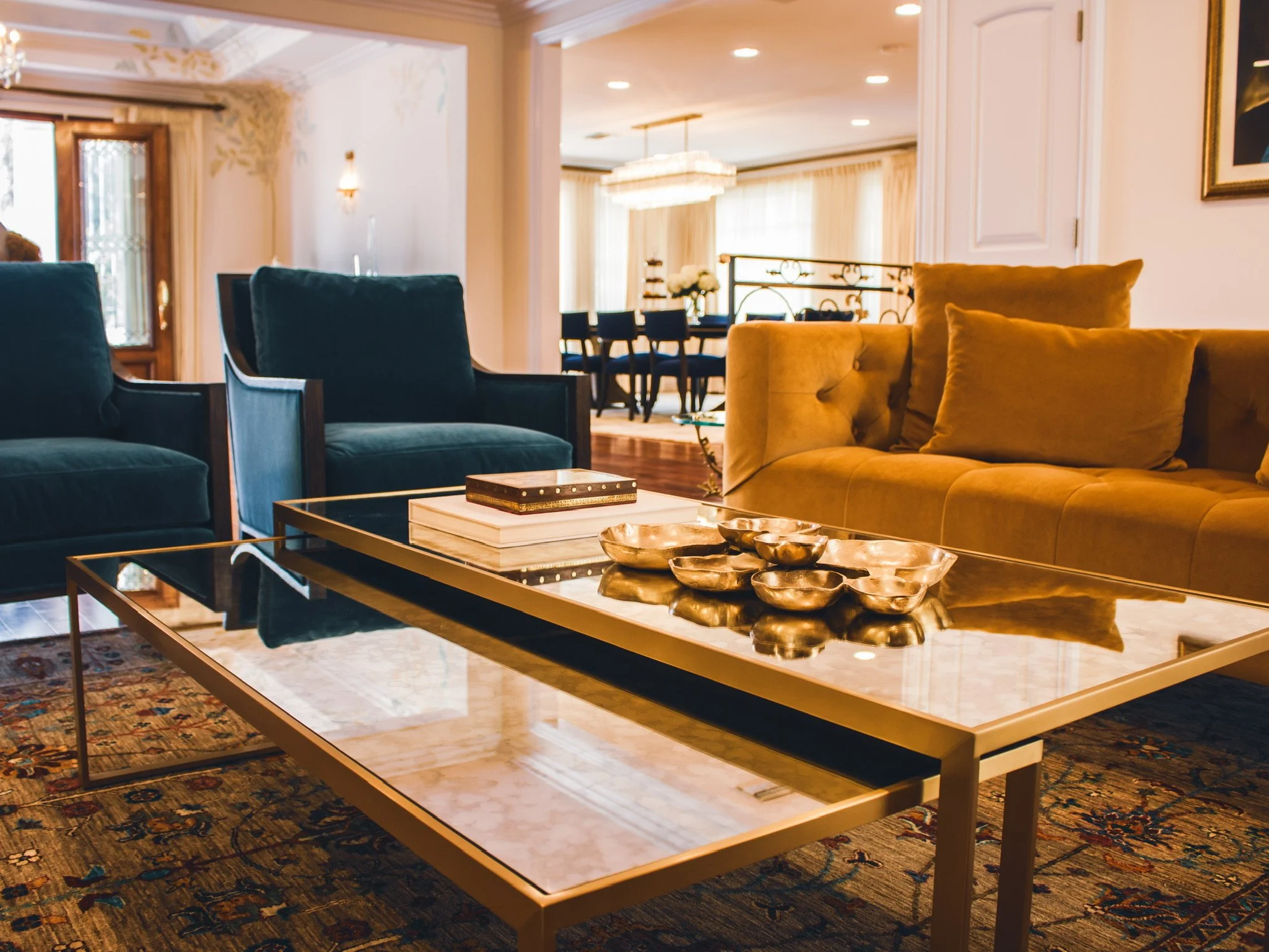 Living room with gold couch, teal armchairs, glass coffee table with decorative bowls, and a dining area in the background.