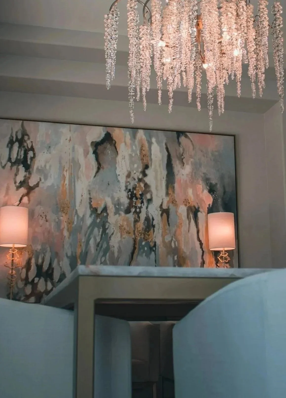 A living room corner with a large abstract painting on the wall, two matching lamps on side tables, and a chandelier with hanging pink crystals overhead.