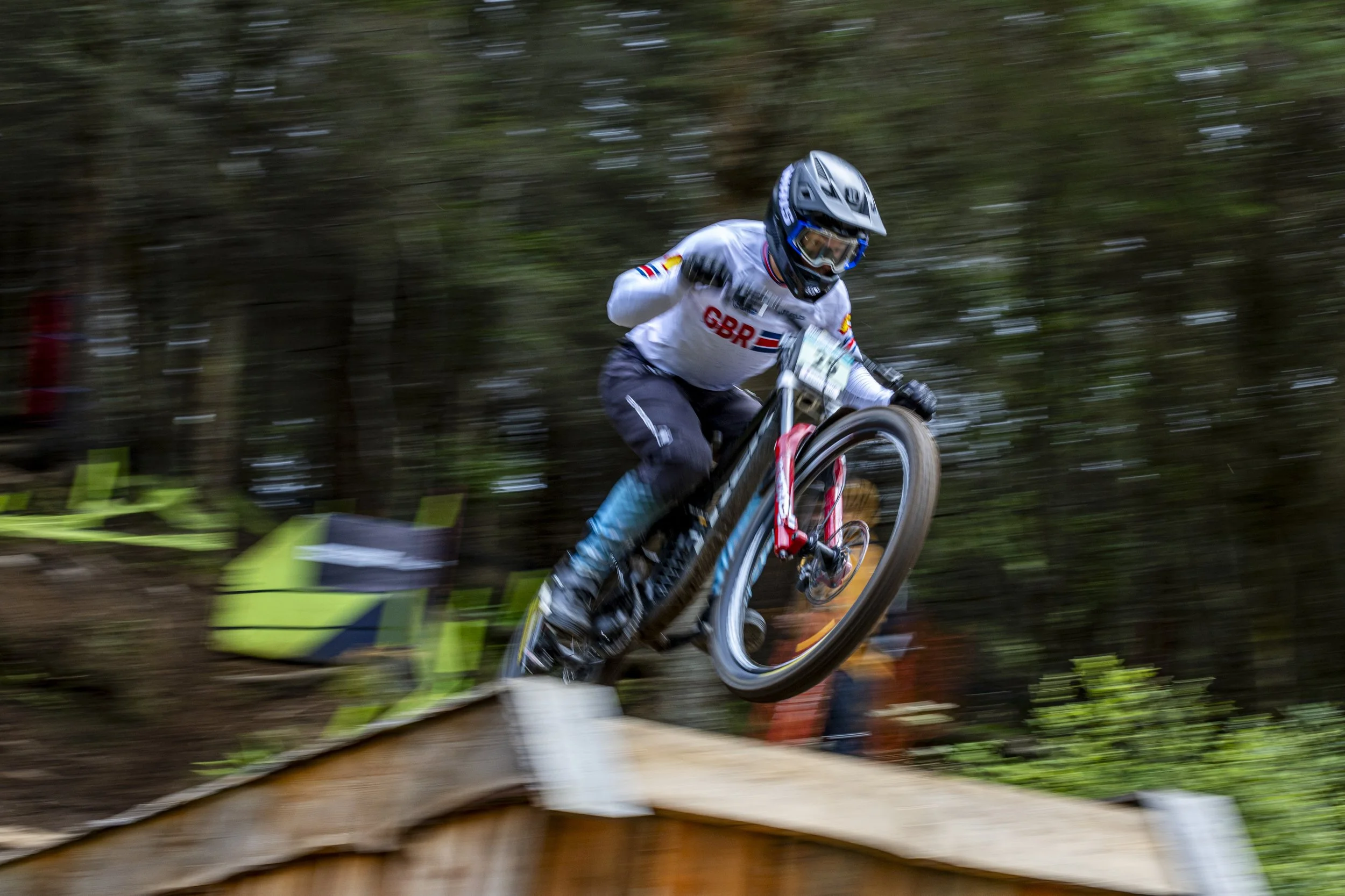 Mountain biker jumping off a wooden ramp in a forested area, wearing a helmet and protective gear.