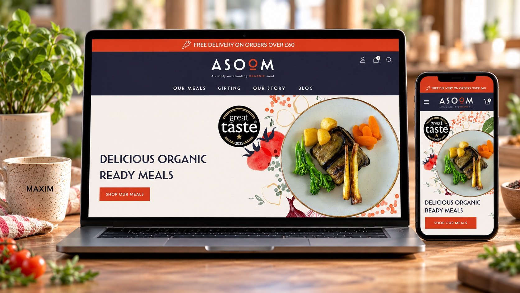 A laptop and smartphone displaying a website for organic ready meals, featuring a plate of grilled vegetables, carrots, and potatoes, on a wooden table with a sprig of cherry tomatoes, a mug with 'MAXIM' printed, and potted plants in the background.