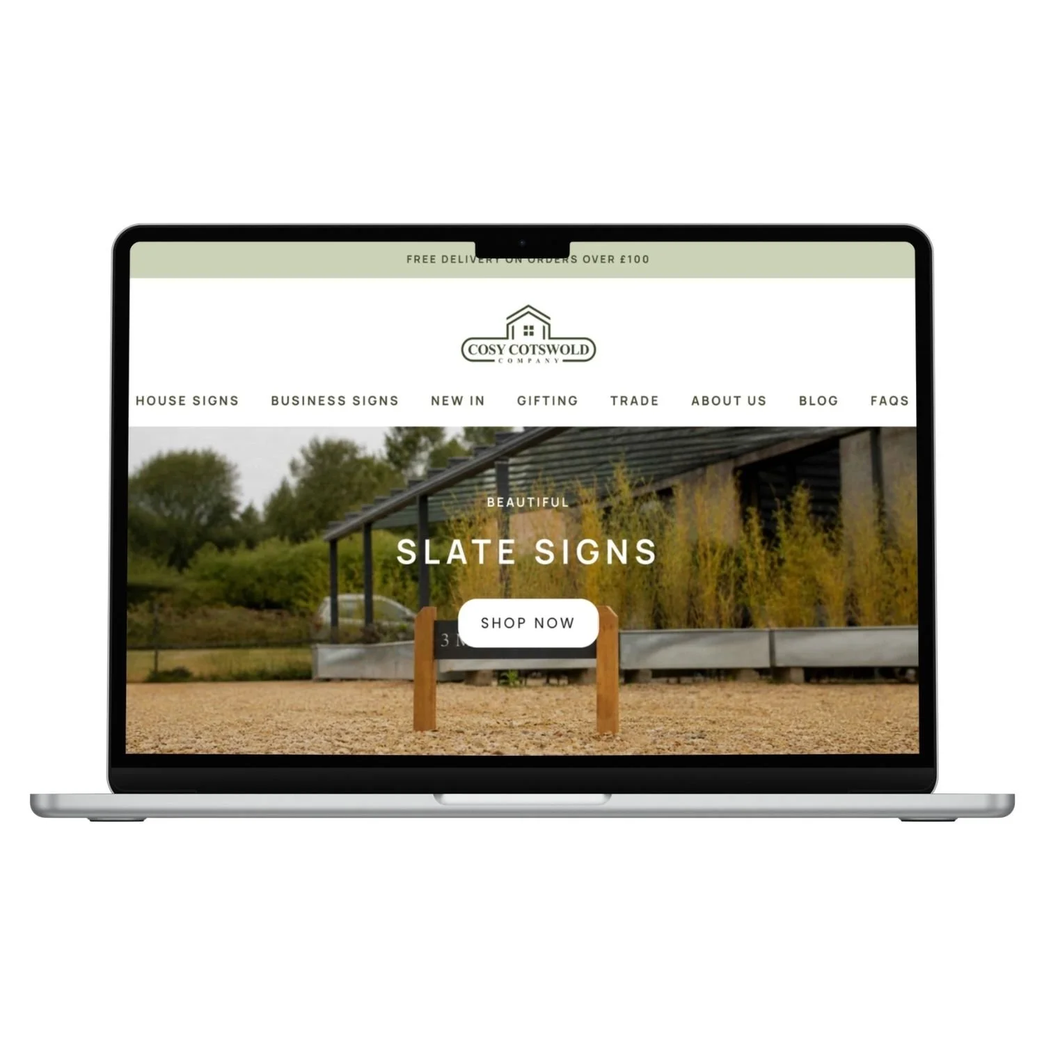 Laptop displaying a website for Cosy Cotswold Company with a header about free delivery on orders over £100, navigation links, a photo of a signs with a background of trees and a building, and a "Shop Now" button for slate signs.