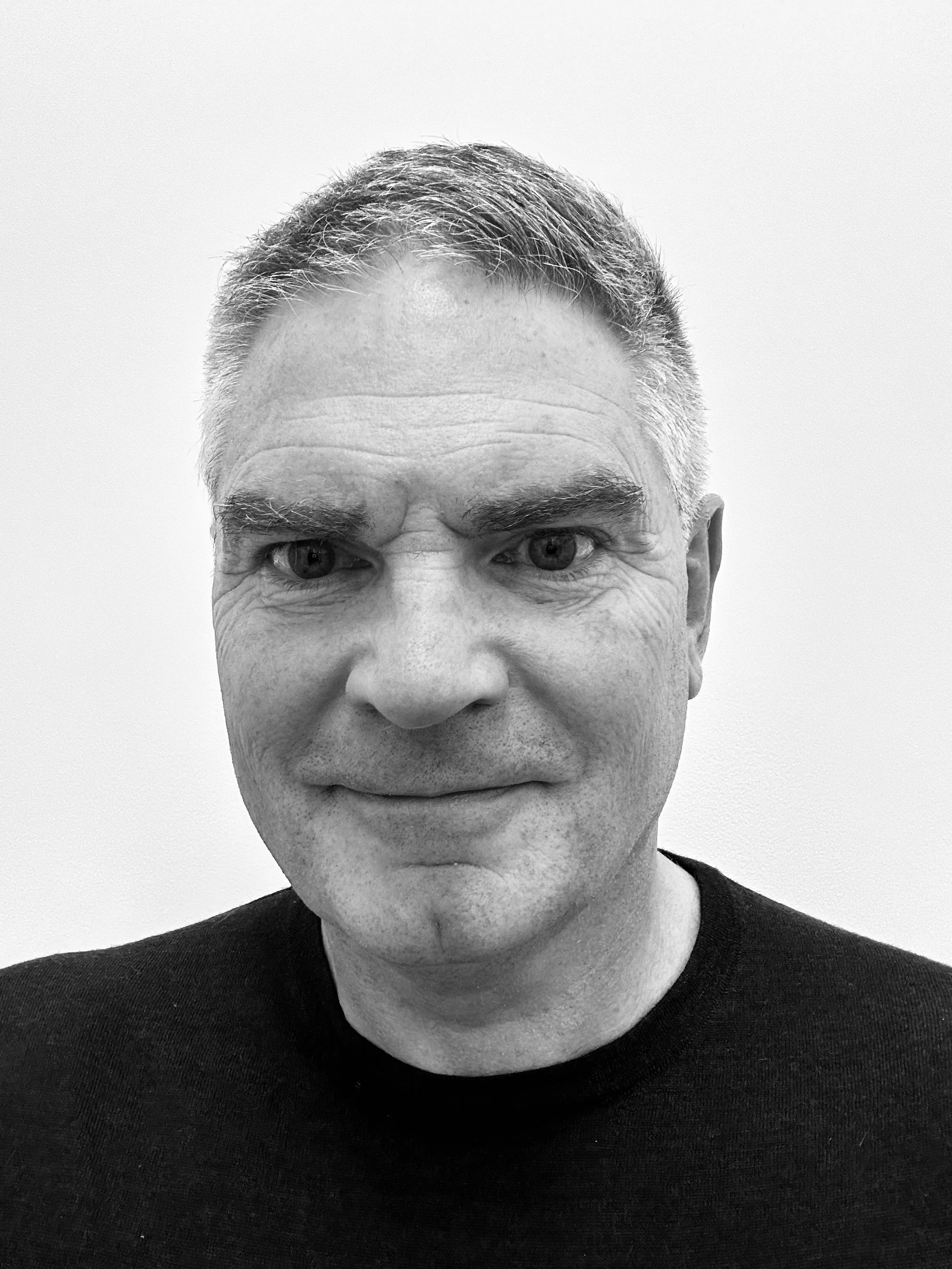 A black and white close-up of a middle-aged man's face with short, silver hair and expressive eyes, wearing a dark shirt against a plain light background.