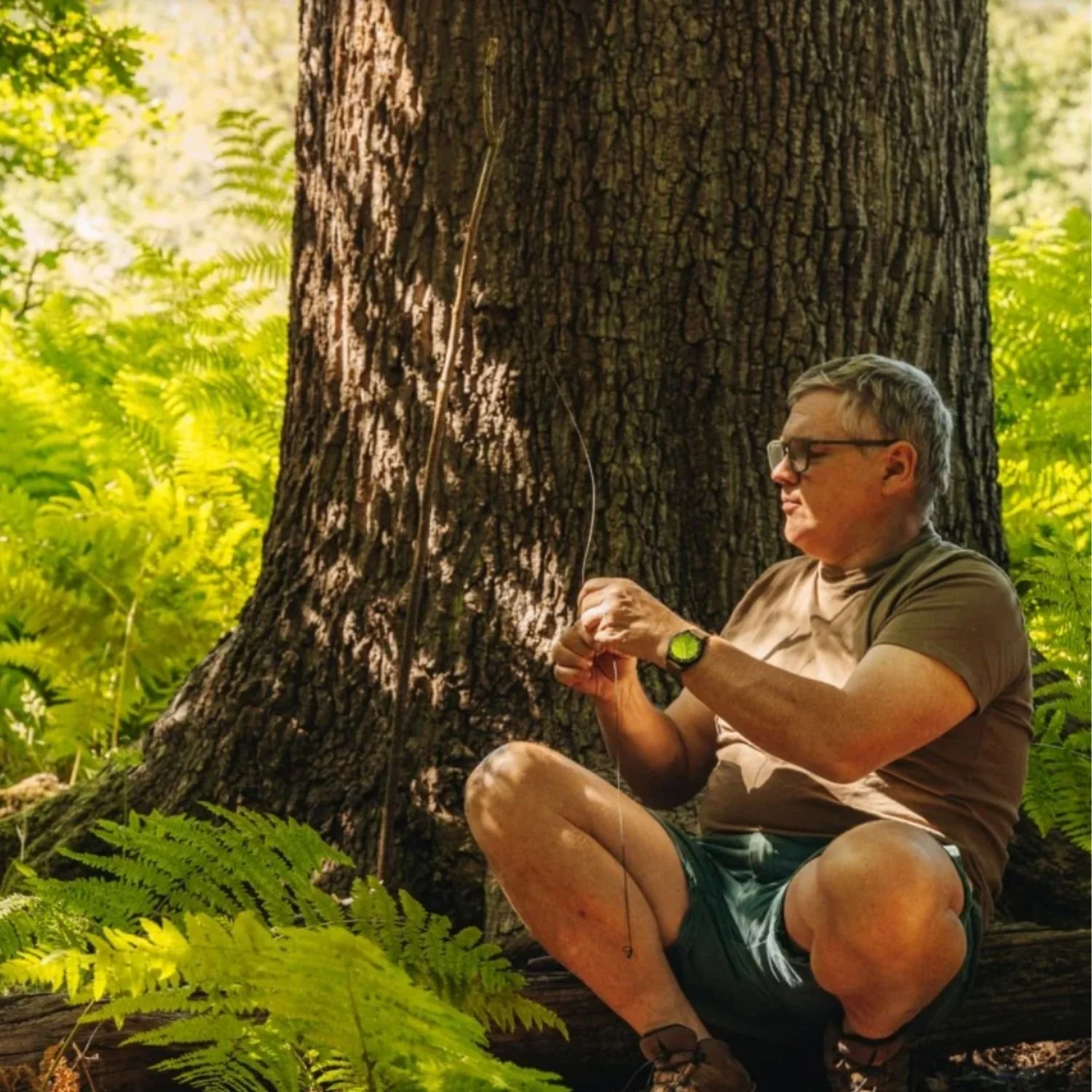 A man with glasses and a watch, sitting outdoors near a large tree, preparing to fish in a lush green forest environment.
