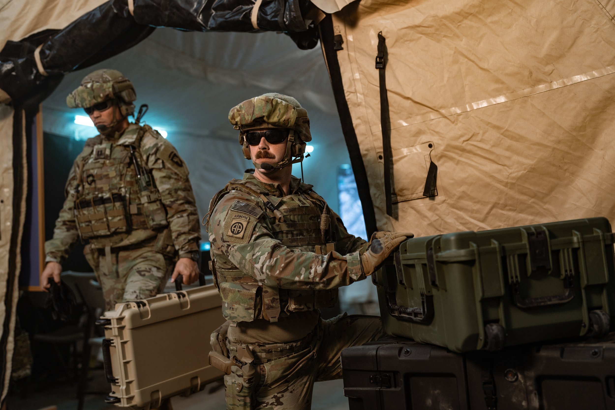 Two soldiers in camouflage uniform with tactical gear, including helmets and sunglasses, are inside a large military tent with beige and black fabric walls. One soldier is standing and the other is kneeling, handling military equipment.