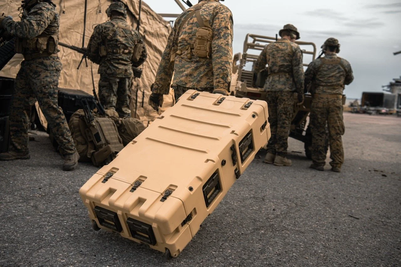 Military personnel in camouflage uniforms working near tents and equipment at an airfield, with one soldier carrying a beige protective case.