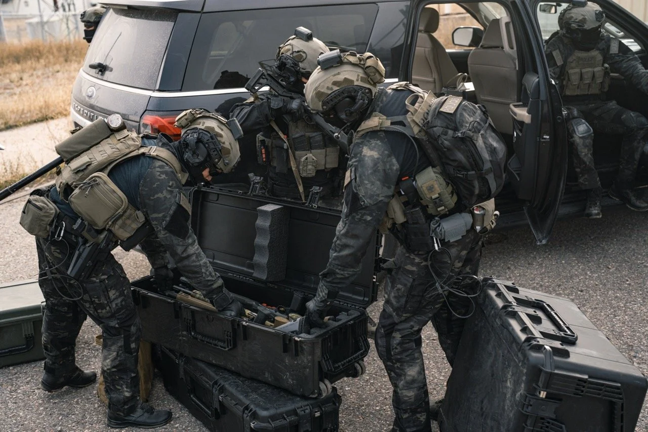 Three military personnel in tactical gear loading equipment from a black case into a vehicle