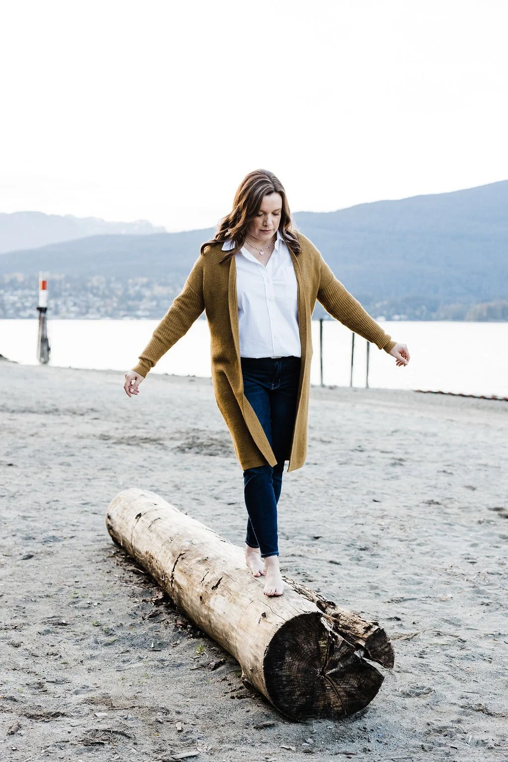 Woman walking barefoot on a log at the beach with water and mountains in the background.