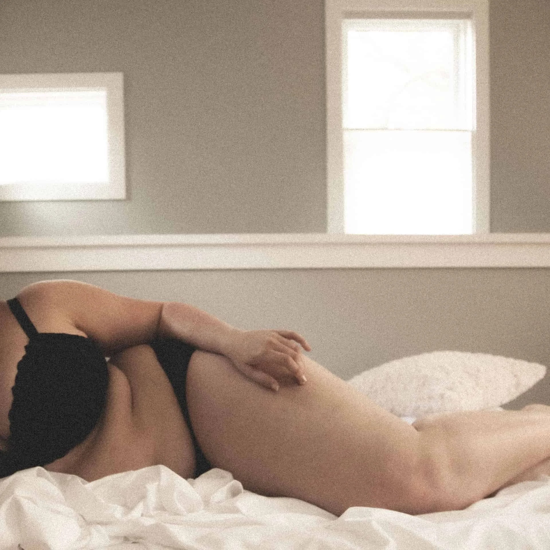 A person lying on their side on a bed, with one hand resting on their thigh, in a bedroom with two windows in the background.