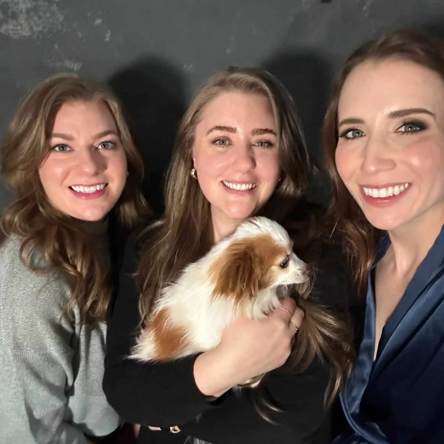 Three women smiling and posing for a photo with a small dog, with a dark background.