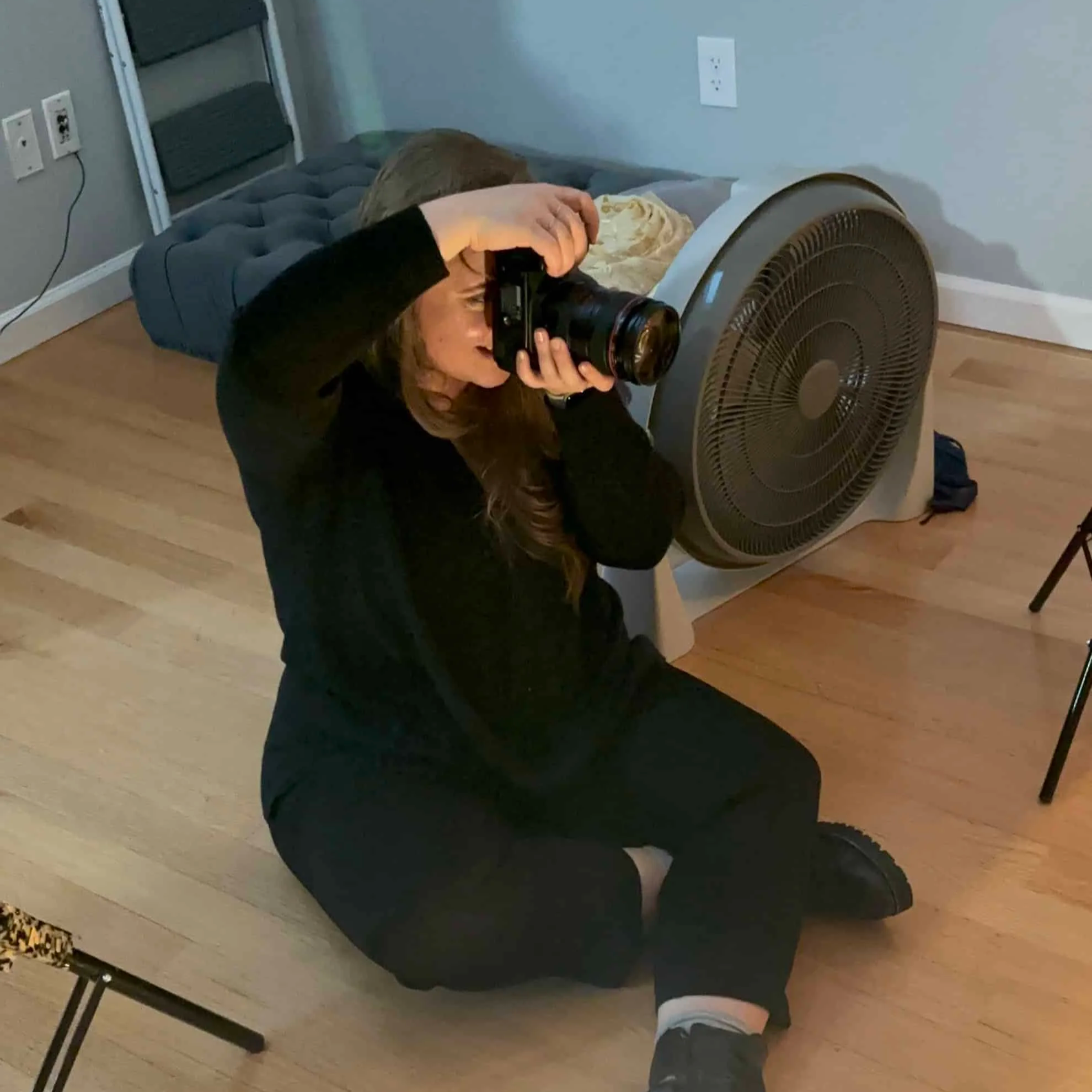 A woman kneeling on a wooden floor, taking a photograph with a camera. She is dressed in black and has long brown hair. There is a large electric fan and a dog bed with a gray cushion against the wall behind her.
