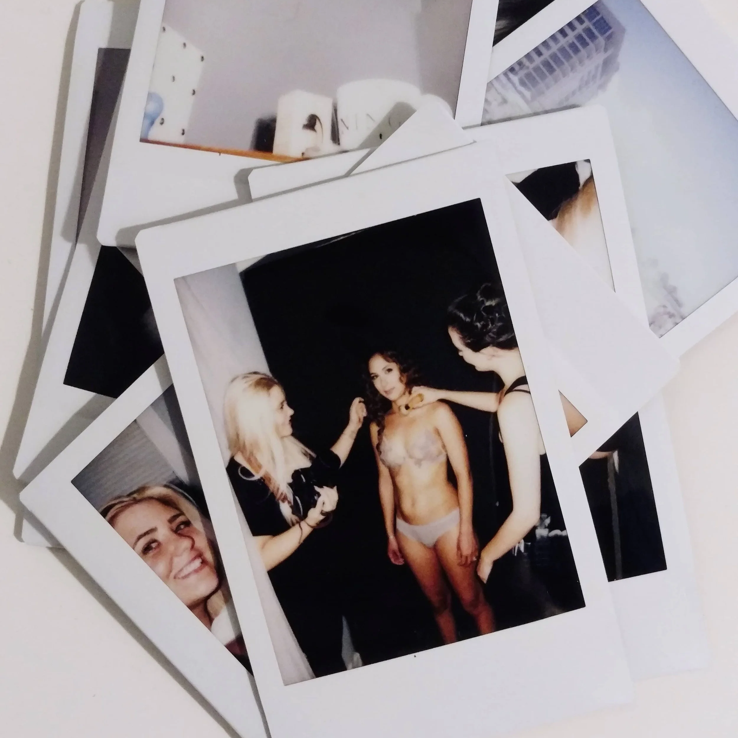A pile of Polaroid photographs on a white surface, with one showing three women, one of whom is partially undressed, in a photoshoot or backstage setting.