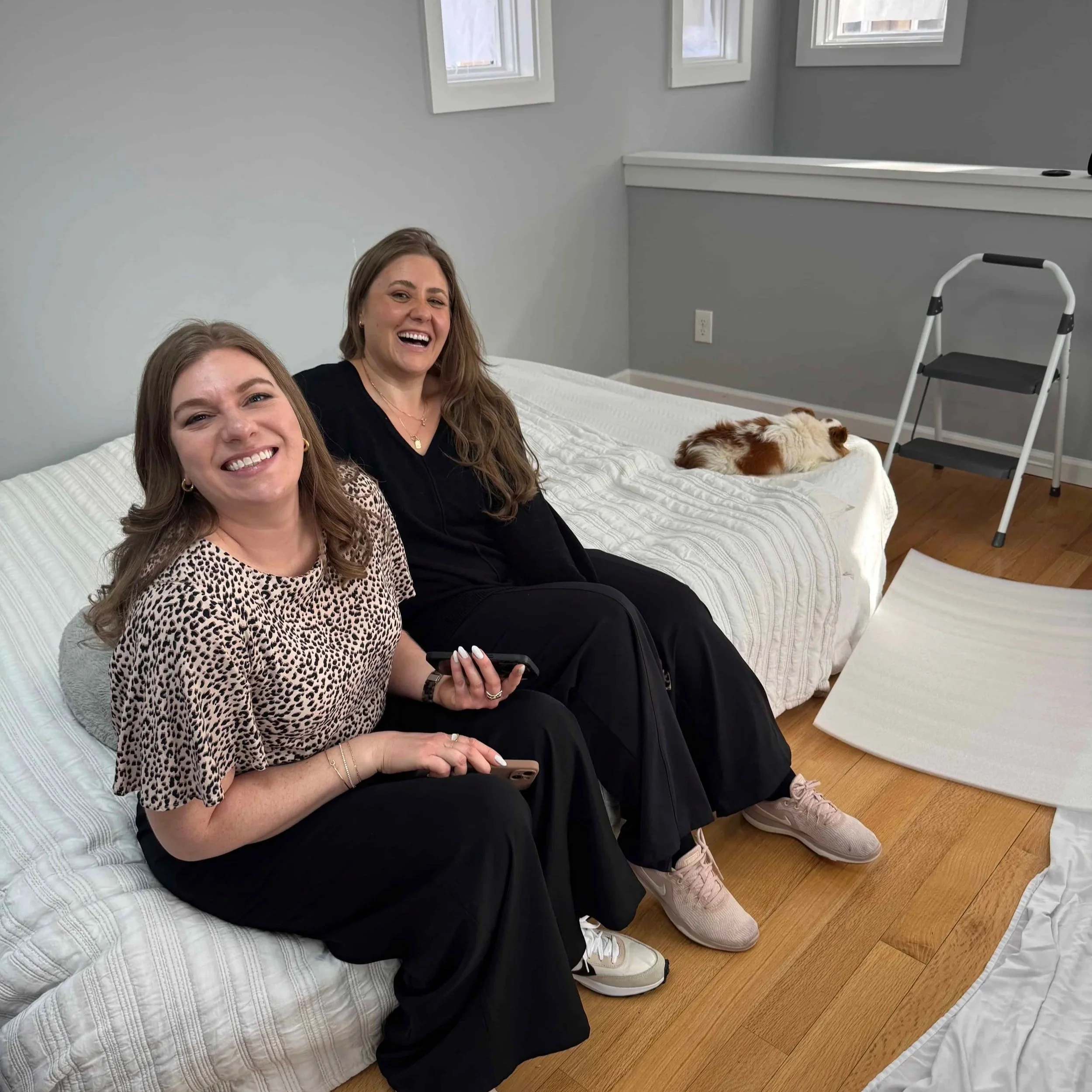 Two women sitting on a bed with a small dog lying beside them. The room has three small windows on a gray wall.