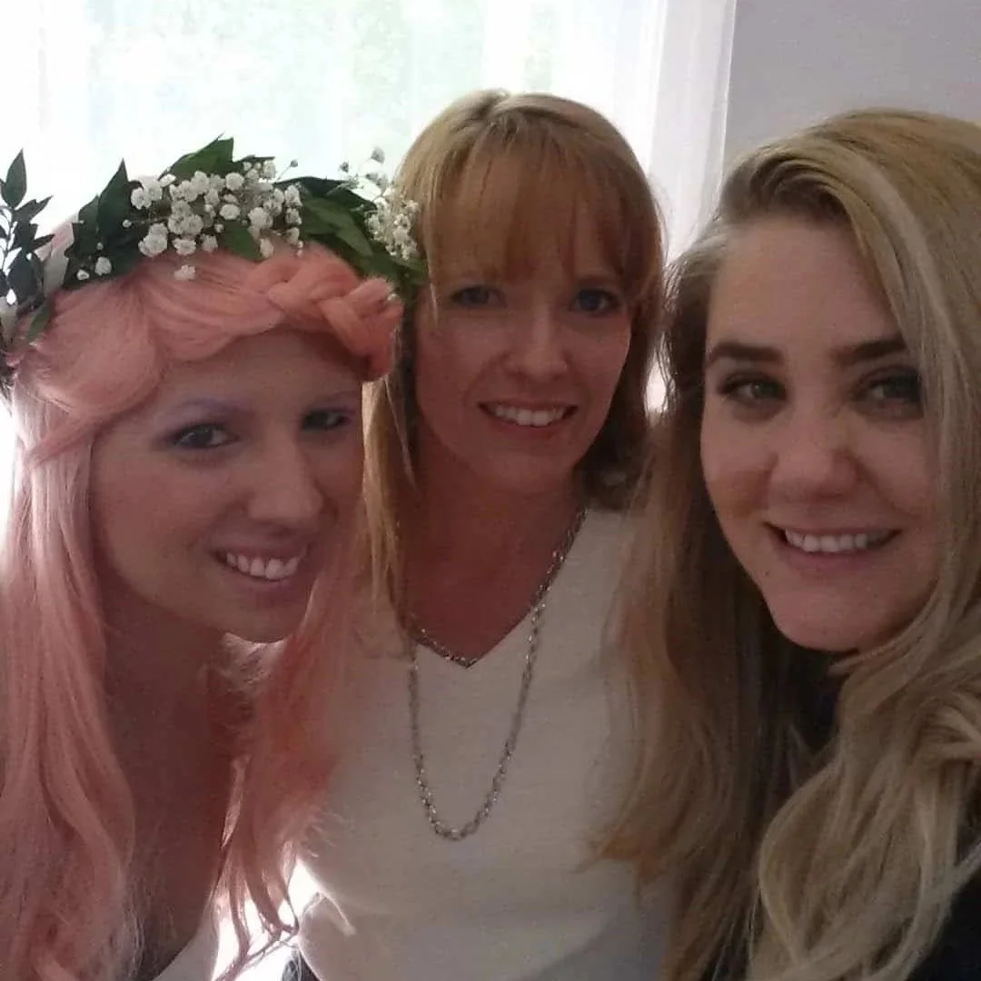 Three women smiling for a selfie, one with pink hair and a floral headband, another with red hair, and a third with blonde hair, in a well-lit room.