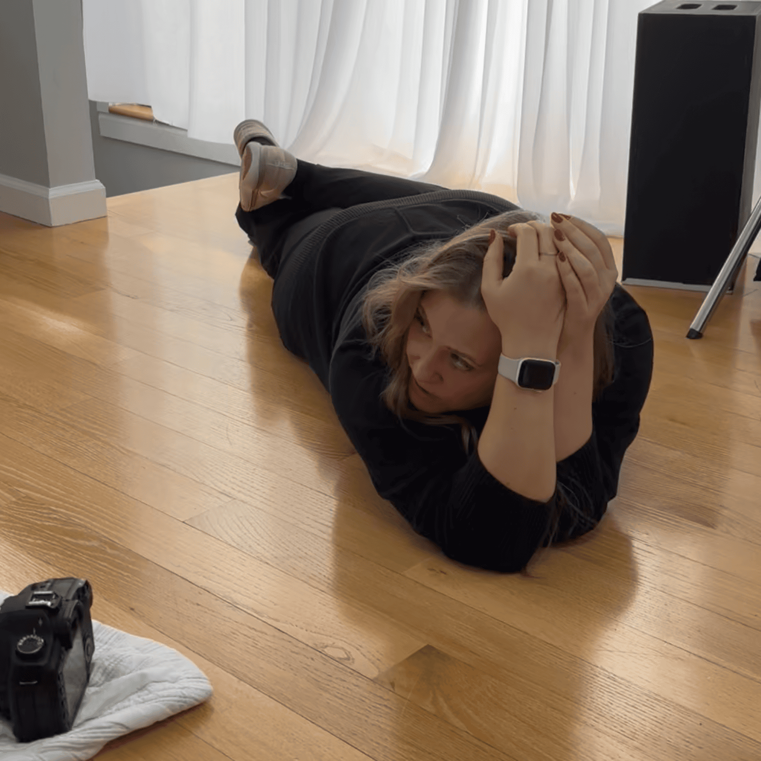 Woman lying on hardwood floor in a plank position, holding her head with both hands, with a camera and a camera strap on a white cloth nearby, and a speaker and a folded tripod in the background.