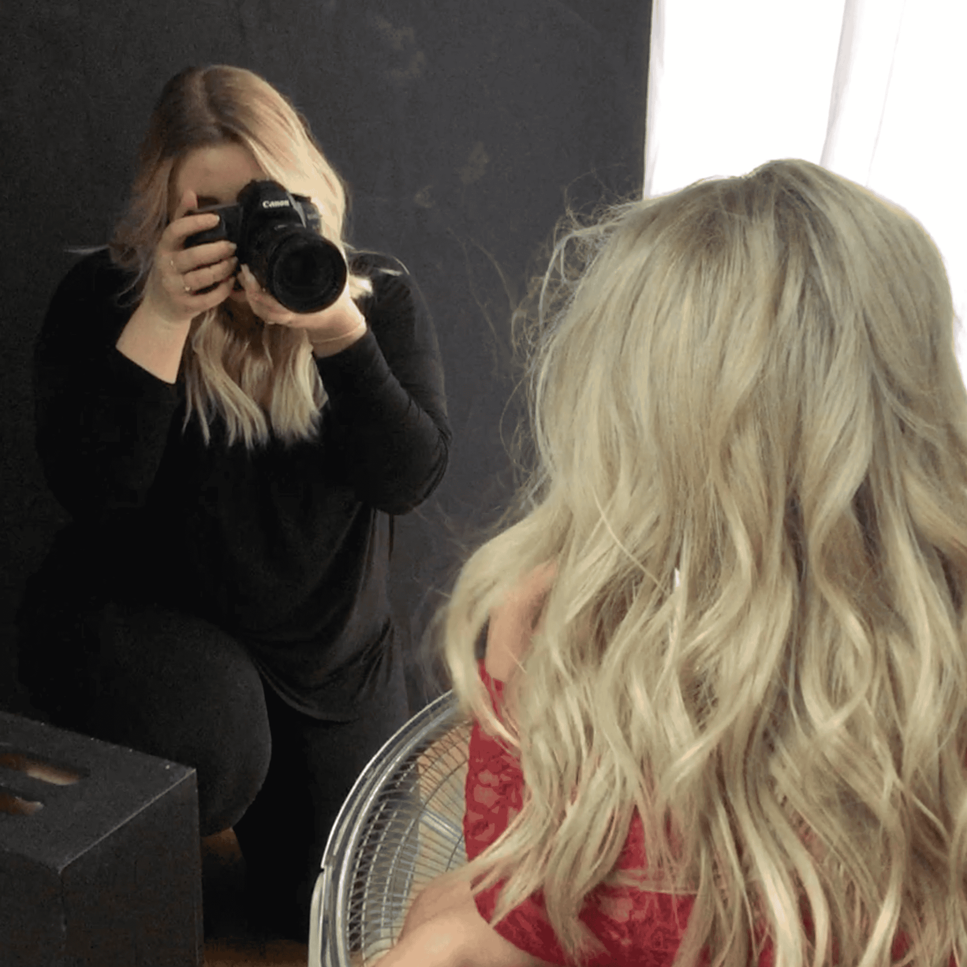Photographer taking a picture of a woman with long, curly blonde hair, seen from behind, sitting in front of a fan, in a studio with black wall and light coming from a window.