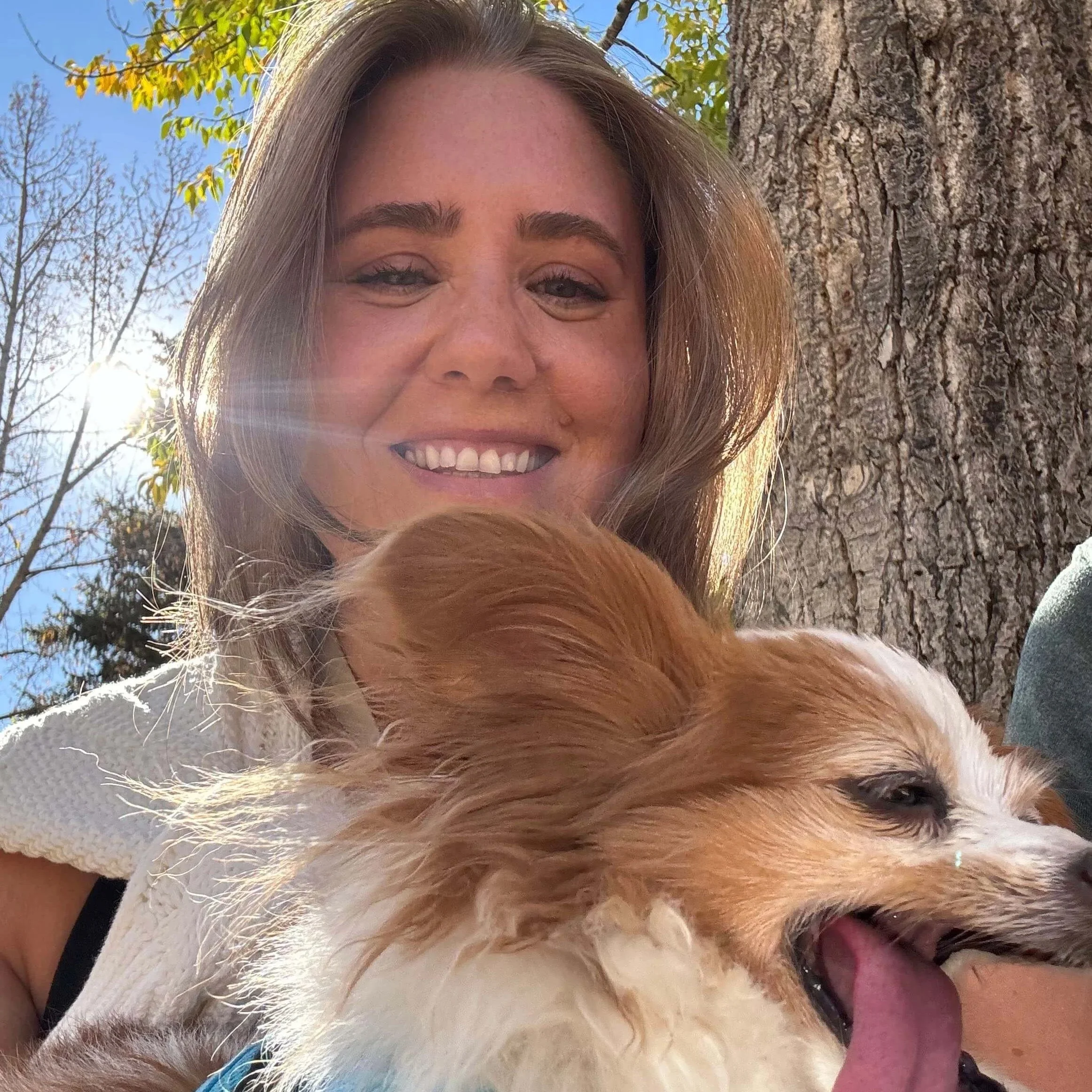 A woman smiling outdoors with a dog, sunlight filtering through the trees in the background.