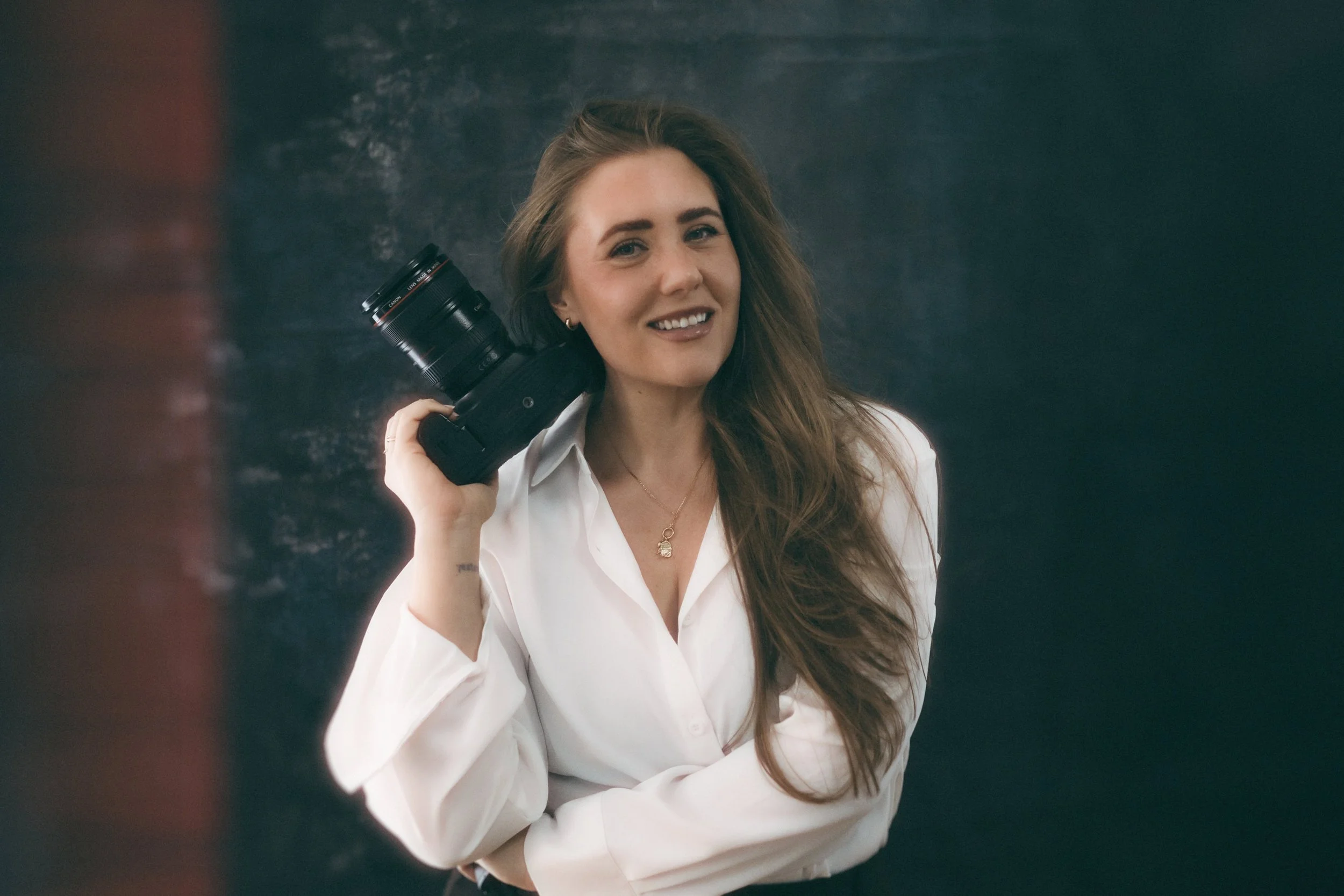 A woman with long brown hair wearing a white shirt, smiling, holding a camera on her shoulder, standing against a dark background.
