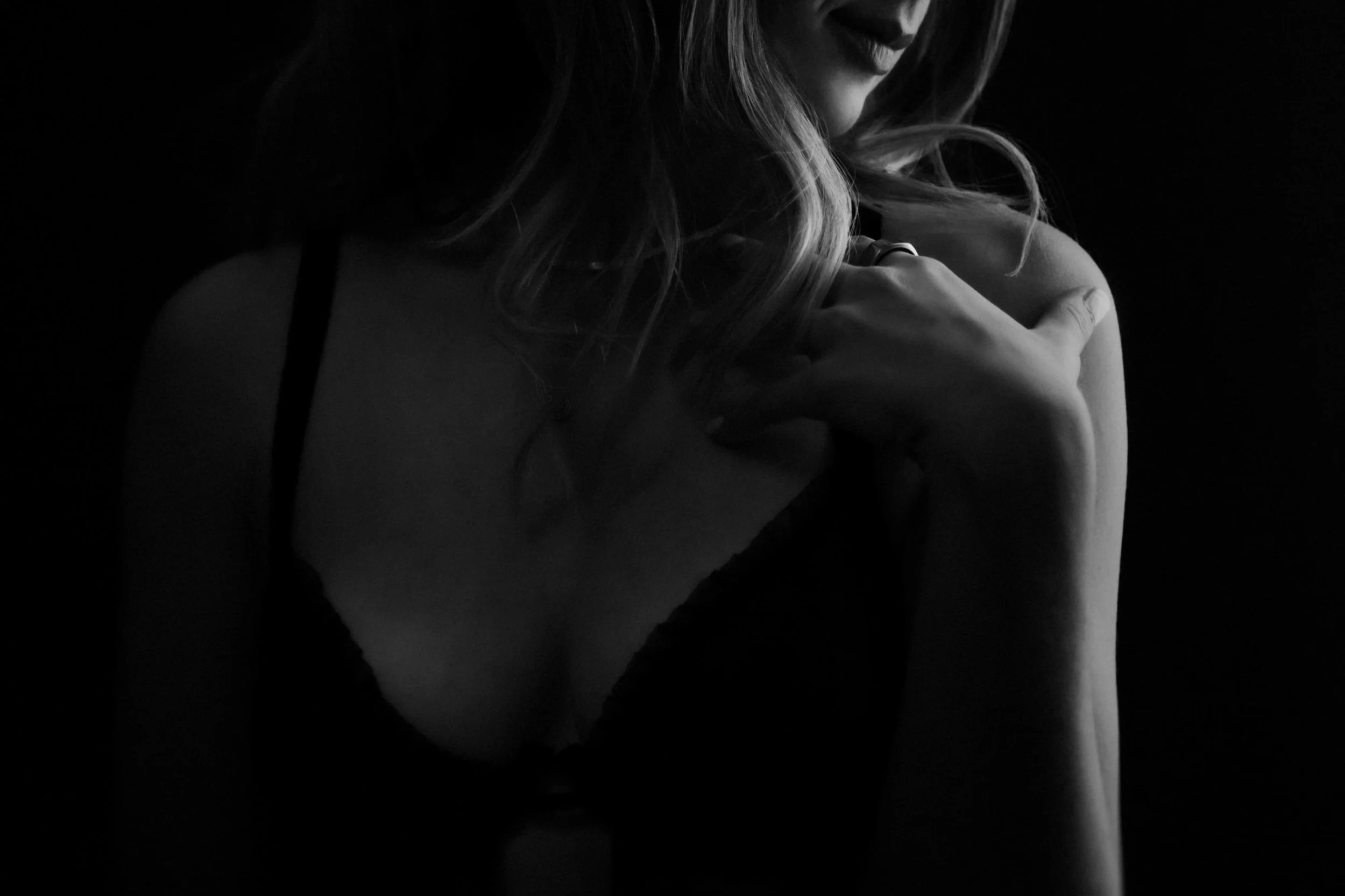 A black-and-white photograph of a woman with long hair, holding her shoulder with her hand, with shadowy lighting.