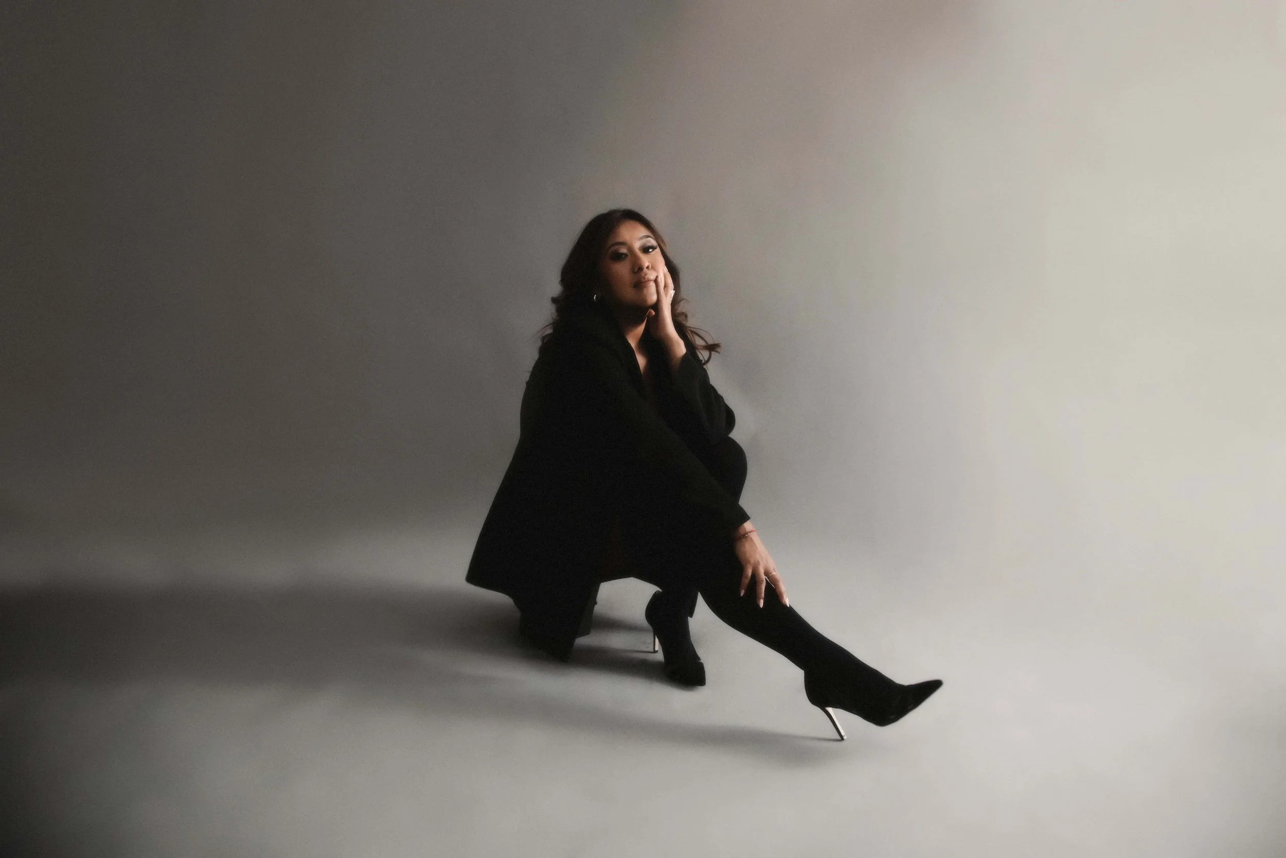 A woman dressed in black high heels and a black coat is seated on a light-colored studio floor against a gray background, with her head resting on her hand and her other hand on her knee.