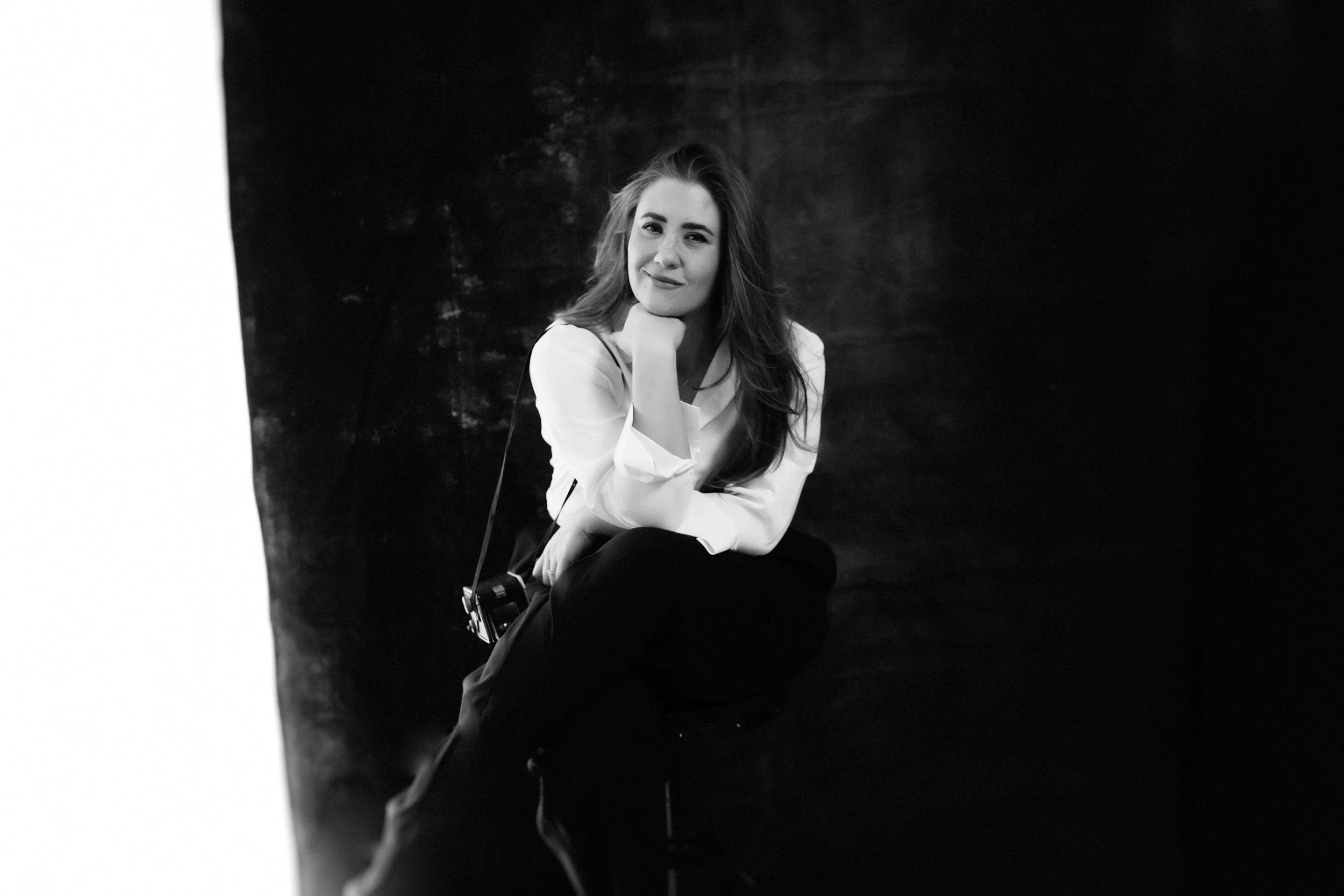 Black and white photo of a woman sitting and smiling, resting her chin on her hand, with a camera hanging from her shoulder, against a dark background.