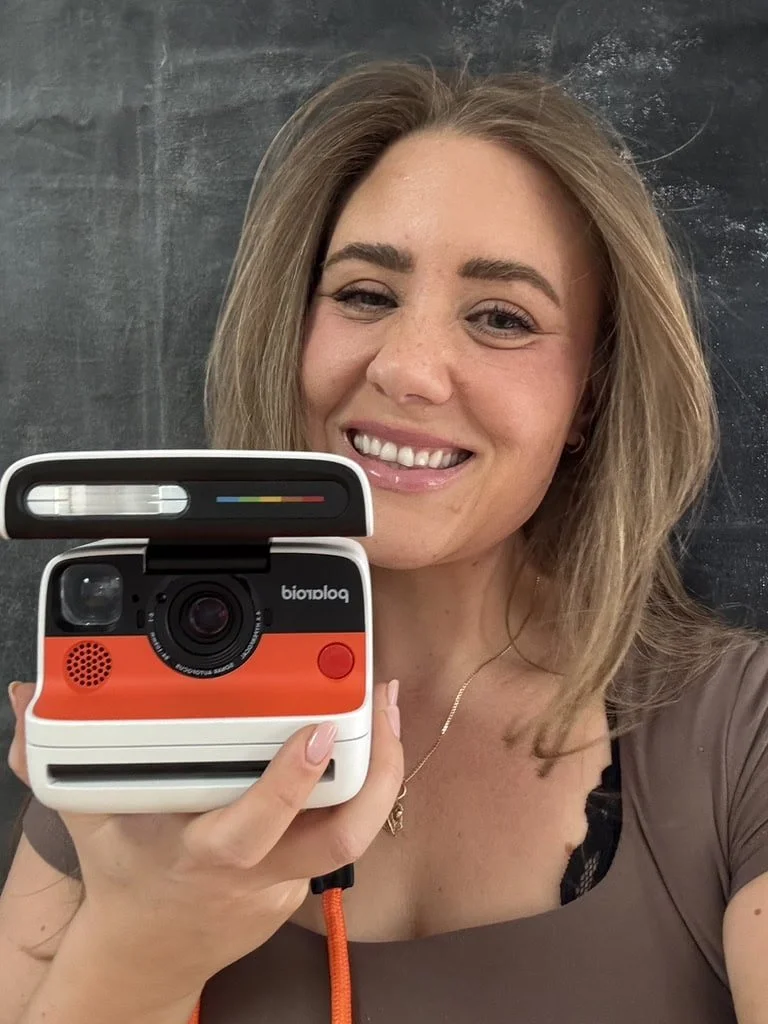 A woman taking a selfie with a Polaroid instant camera, smiling in front of a dark background.