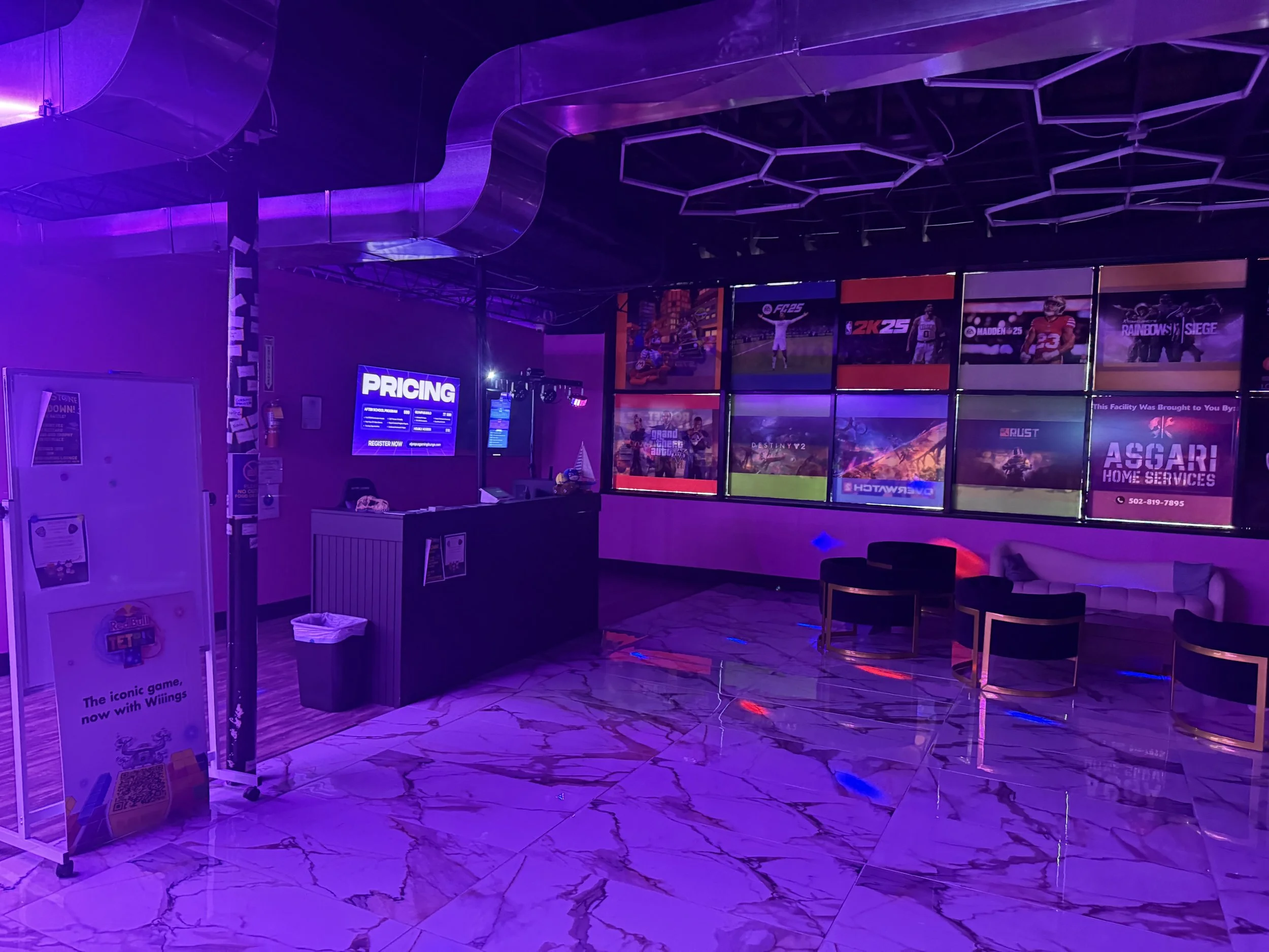 Empty gaming lounge with purple lighting, marble floor, row of monitor screens displaying game advertisements, lounge chairs, and a reception desk with a pricing sign.