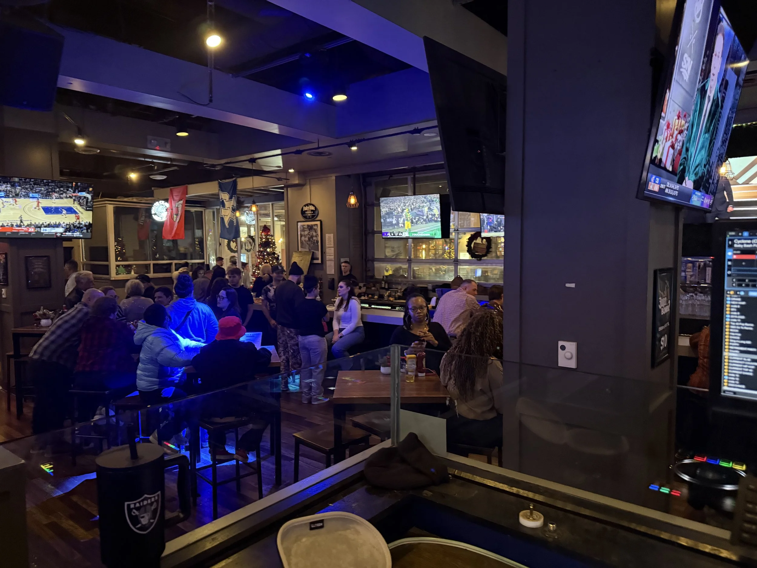 Inside a sports bar filled with people watching multiple television screens showing basketball games. The bar is decorated with flags and a Christmas tree. Some patrons are seated at tables, others standing and socializing. The scene is lively with w