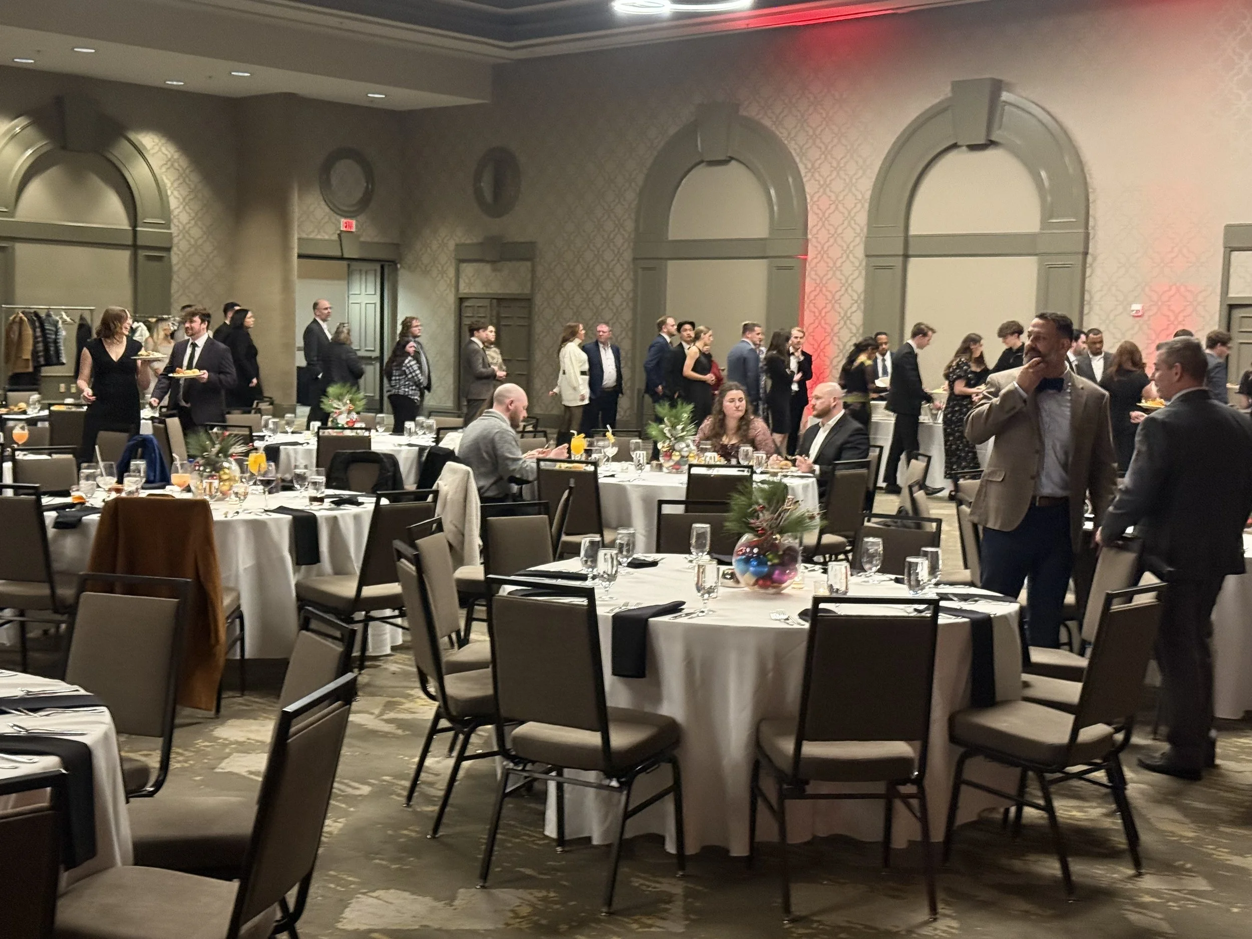 A banquet hall set up for a formal event with round tables decorated with centerpieces and place settings. People are mingling and walking around, some seated and some standing, dressed in business or formal attire.