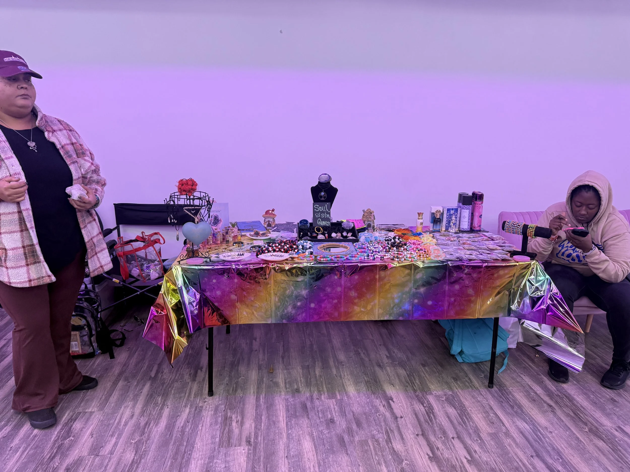 A jewelry and accessory display table with various jewelry pieces, accessories, and small decorative items, set against a plain white wall, with two women nearby—one standing on the left and another sitting on a chair on the right, using a phone.