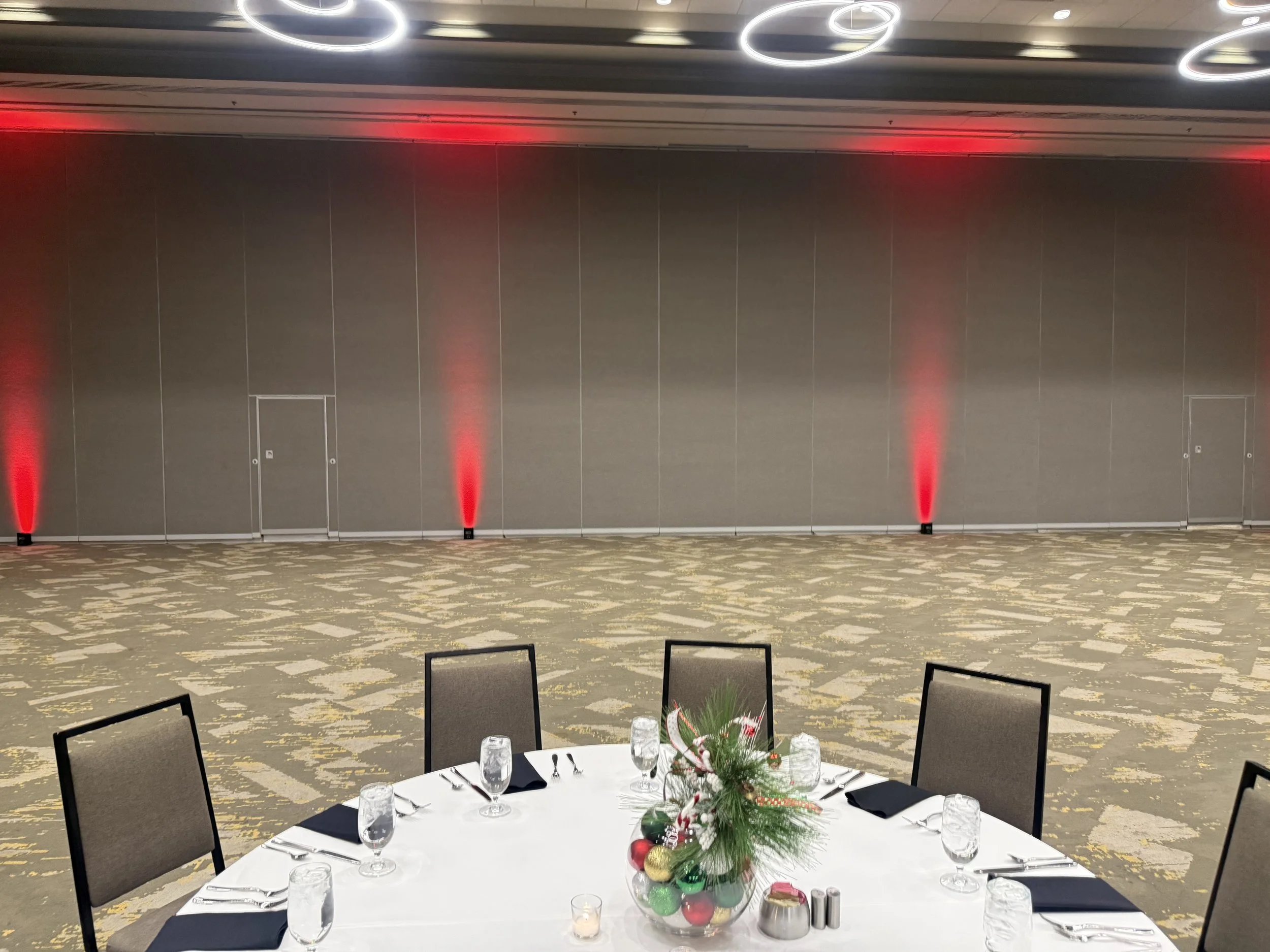 A decorated banquet table set for a meal, with black napkins, glasses, and silverware, in front of a plain wall with red uplighting, with a floral holiday centerpiece and festive ornaments.