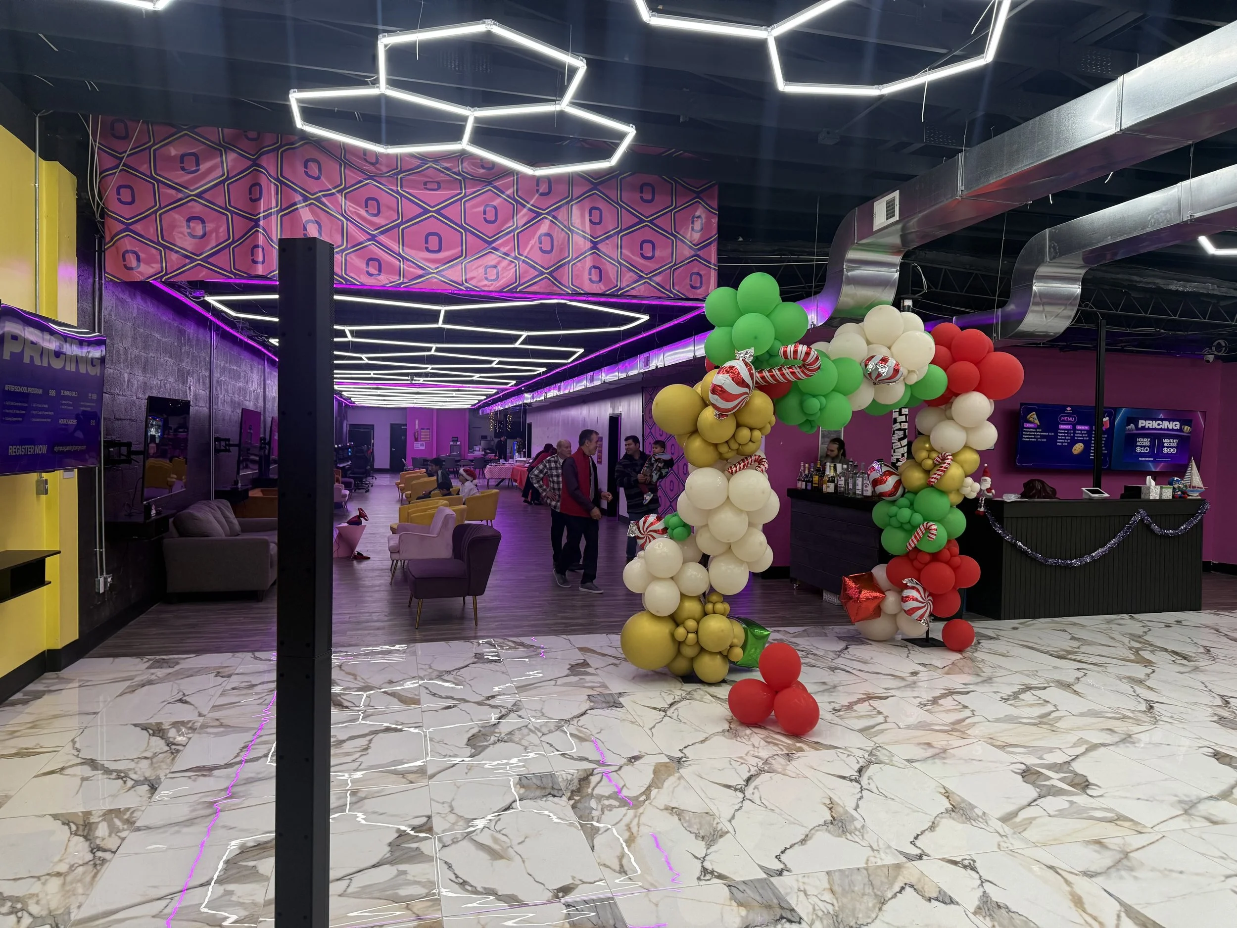 Interior of a modern lounge or club with colorful balloon arch featuring holiday colors and a neon ceiling design, purple walls, a bar area, and seating.