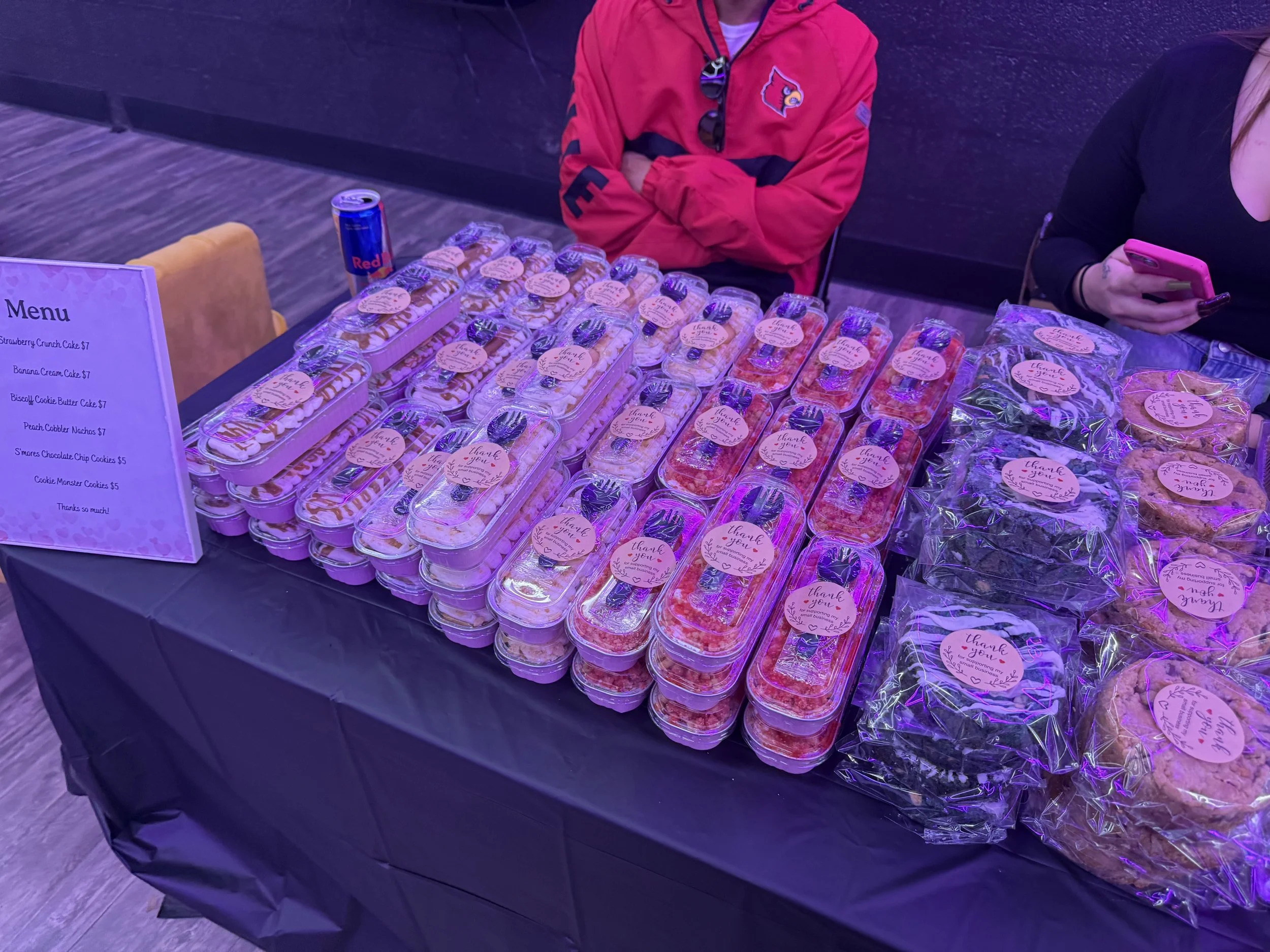 Table filled with assorted decorated cookies in clear containers and plastic bags, with a pink 'Thank You' sticker on each. A purple-lit sign lists cookie flavors and prices, and two women are behind the table, one in a red jacket and the other holdi