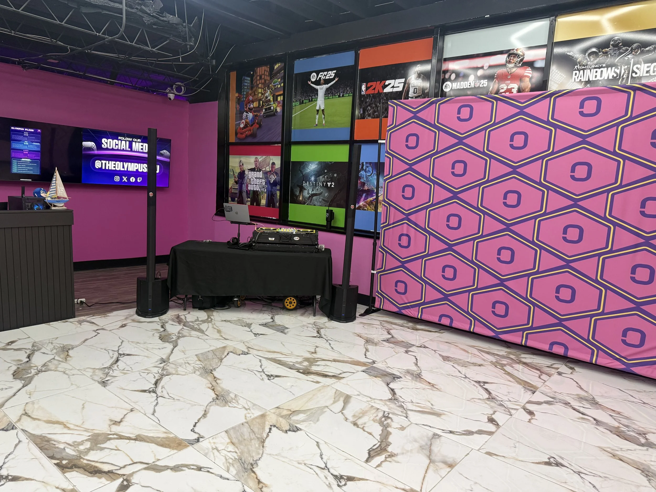 A gaming area with a pink wall, large screen displaying social media information, multiple video game posters on the wall, a pink patterned privacy screen, and a table with DJ equipment.