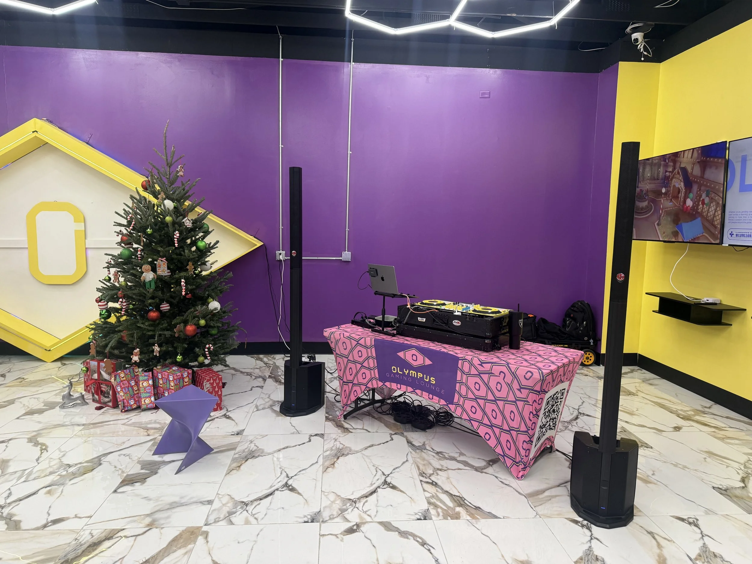 A Christmas tree decorated with ornaments and surrounded by wrapped presents in front of a purple wall with a yellow geometric decoration. To the right, a DJ station with a pink tablecloth, DJ equipment, and speakers, located in a gaming lounge with 