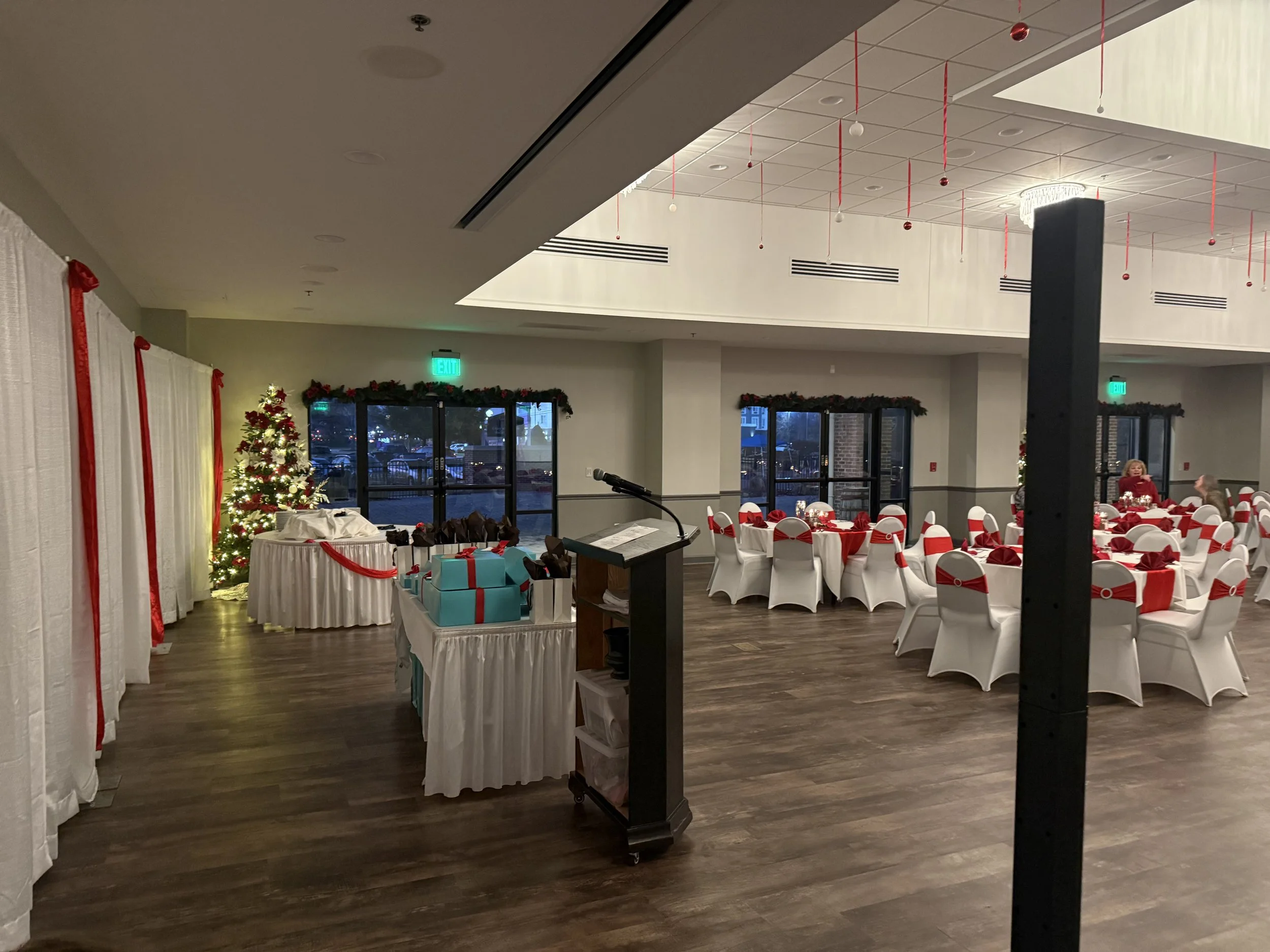 A decorated indoor event space with Christmas decorations, including a lit Christmas tree, wrapped gifts, and tables with red and white table settings, set for a holiday celebration.