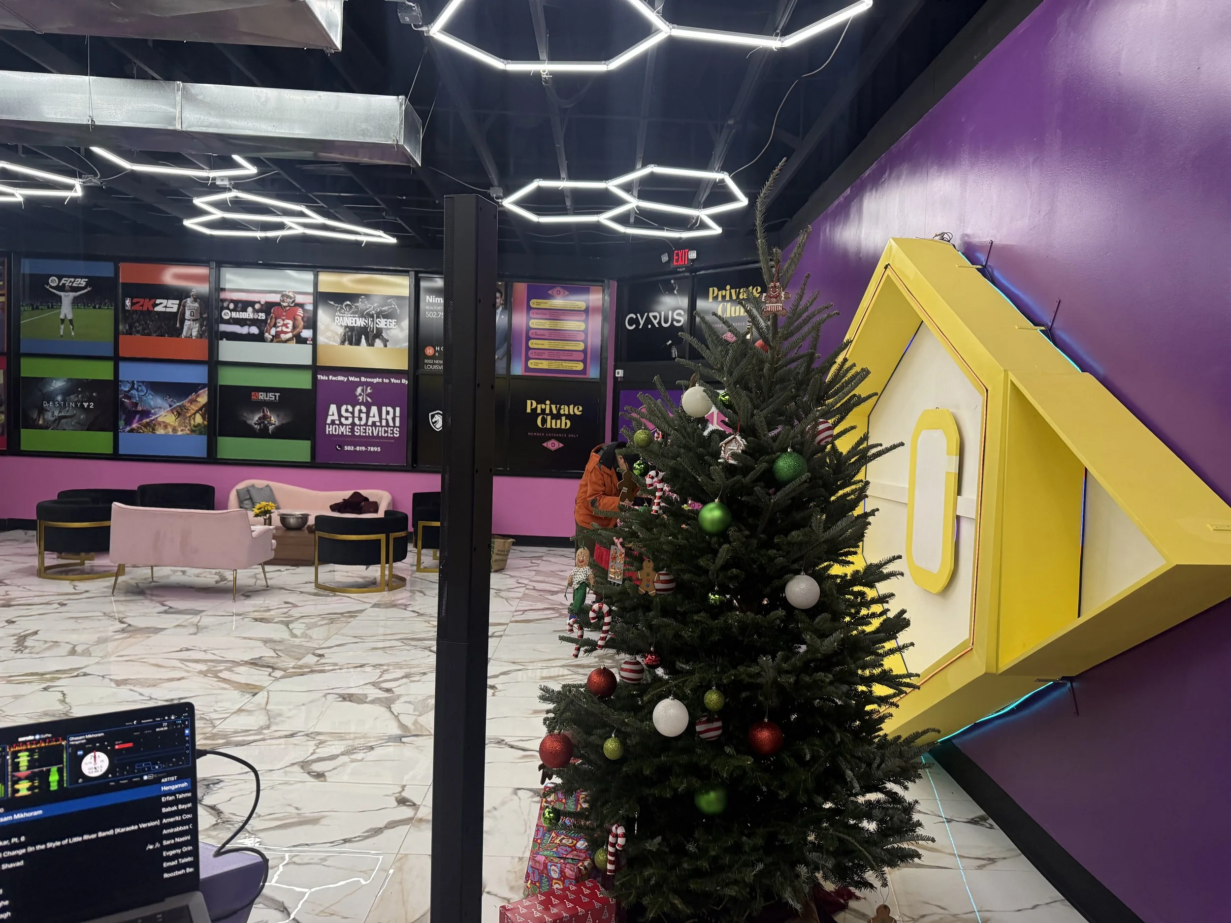 A decorated Christmas tree with red, green, and white ornaments, candy canes, and wrapped gifts underneath, situated inside a colorful lounge or entertainment area with pink, purple, and yellow walls, modern seating, and a wall of gaming posters.