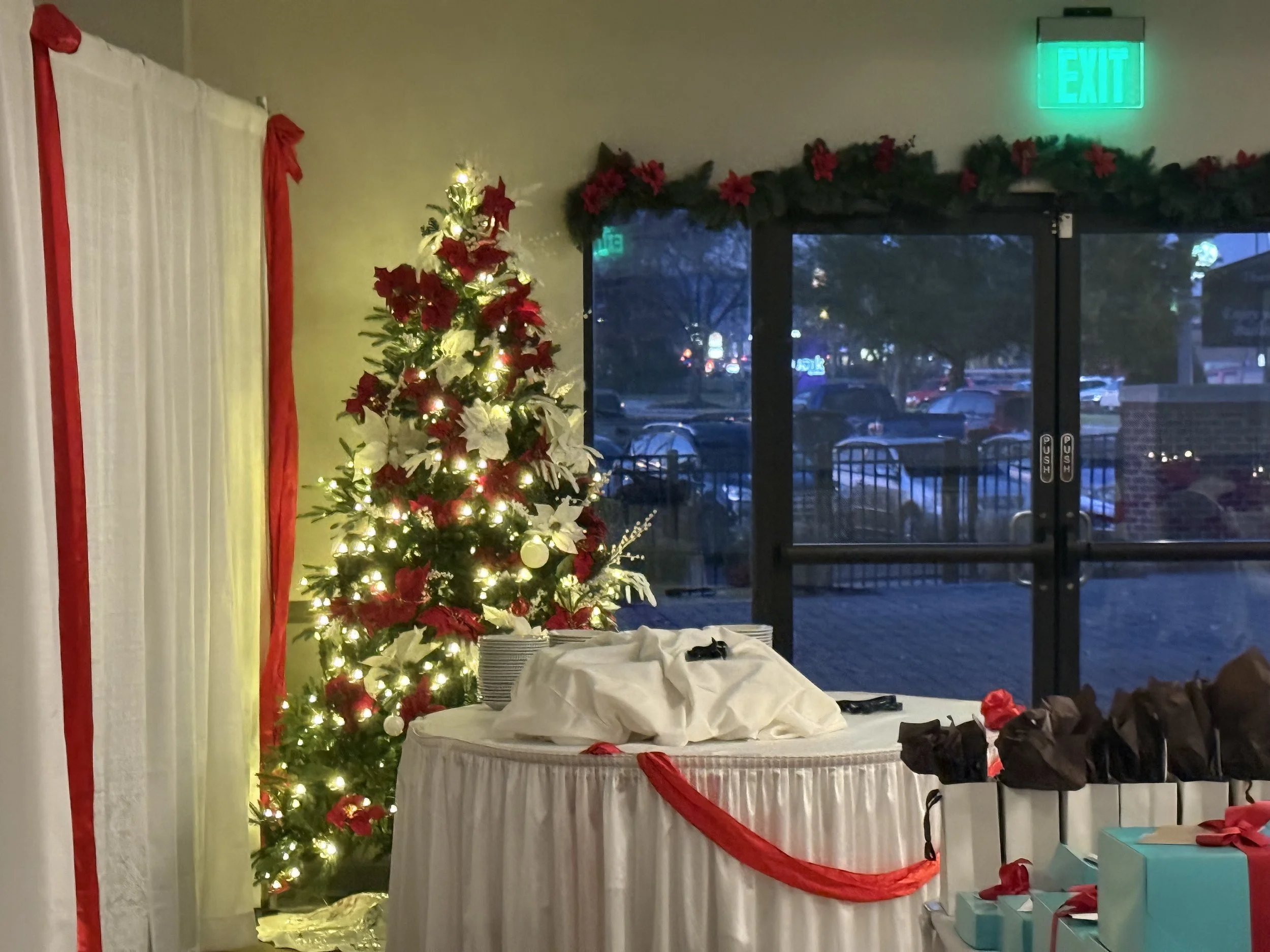 A decorated Christmas tree with white lights and red and white ornaments next to a table covered with a white cloth and red decorations, with wrapped presents nearby. Behind is a glass door with an 