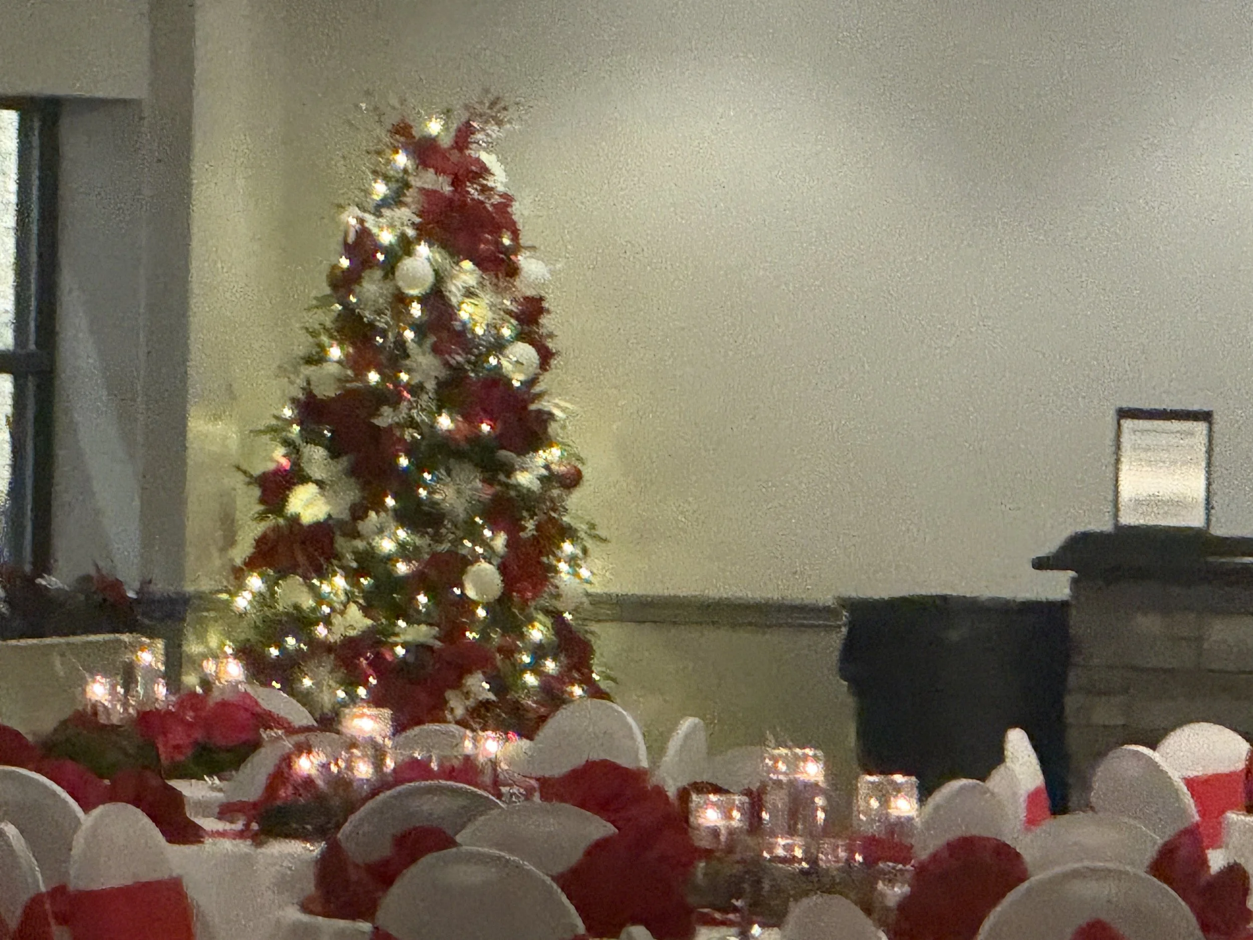 Decorated Christmas tree with white and red ornaments and lights in a banquet hall, surrounded by tables with candlelit centerpieces and white chairs with red ribbons.
