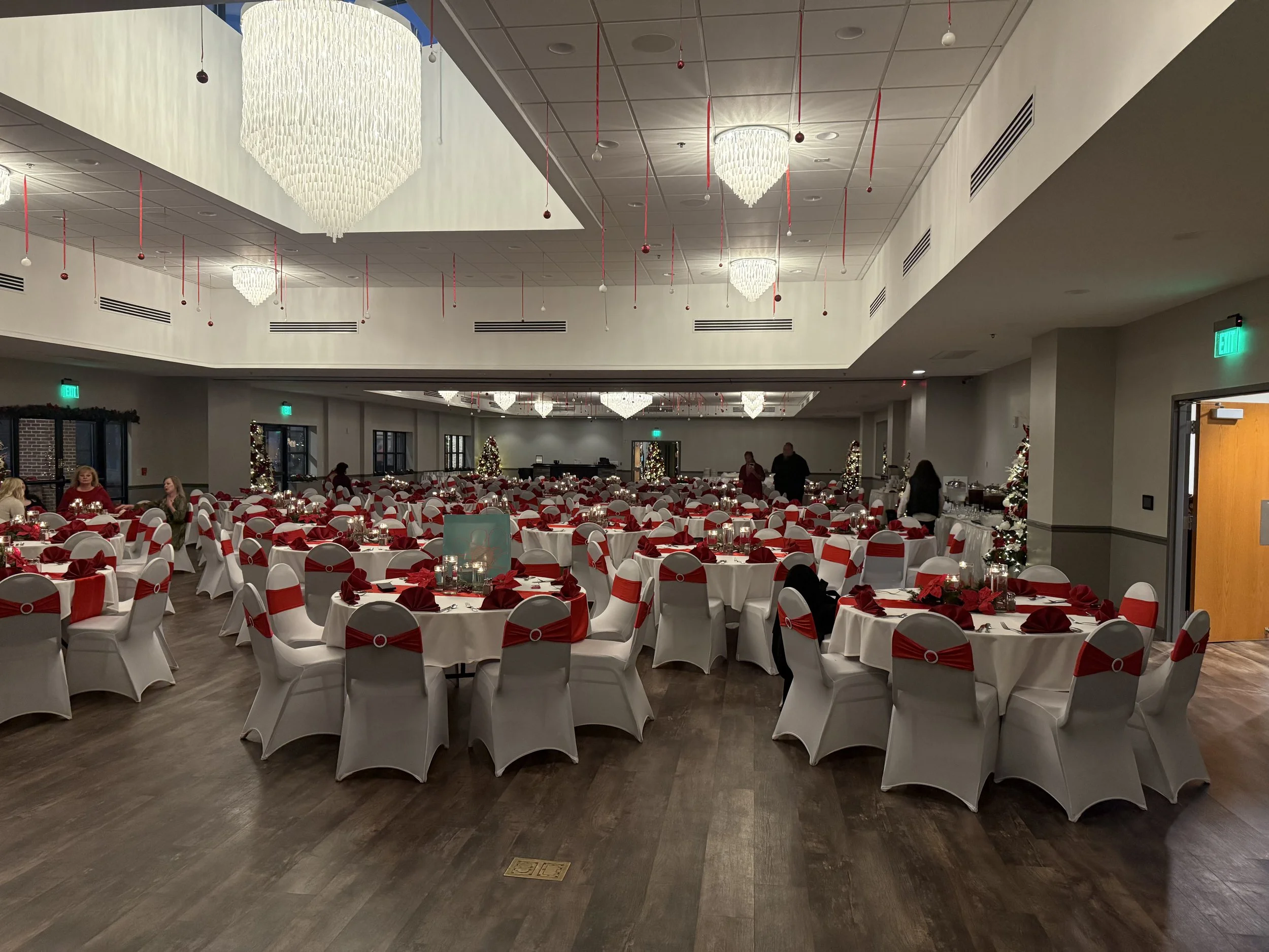 Decorated banquet hall for Christmas with round tables covered in white tablecloths, adorned with red napkins and centerpieces. The room has white chairs with red sashes, Christmas trees, and holiday decorations, under large chandeliers.