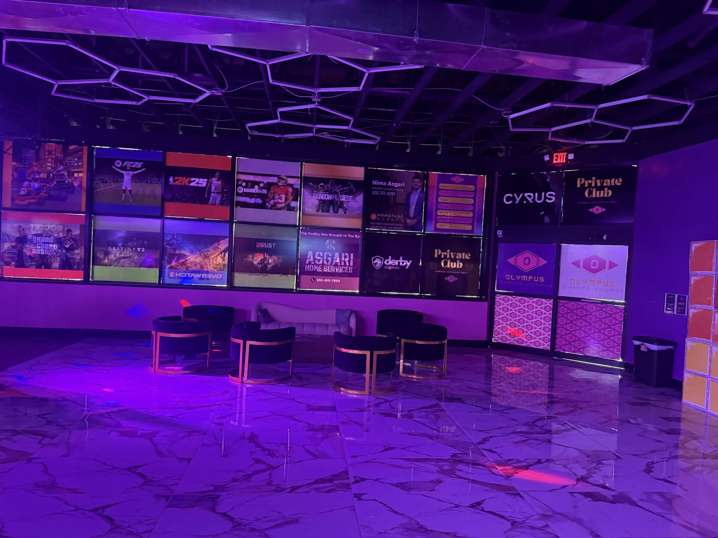 An indoor lounge area illuminated with purple and pink lighting, featuring a white sofa, five black chairs with gold trim, and a marble floor. The back wall has multiple screens displaying various game and entertainment advertisements, with some logo